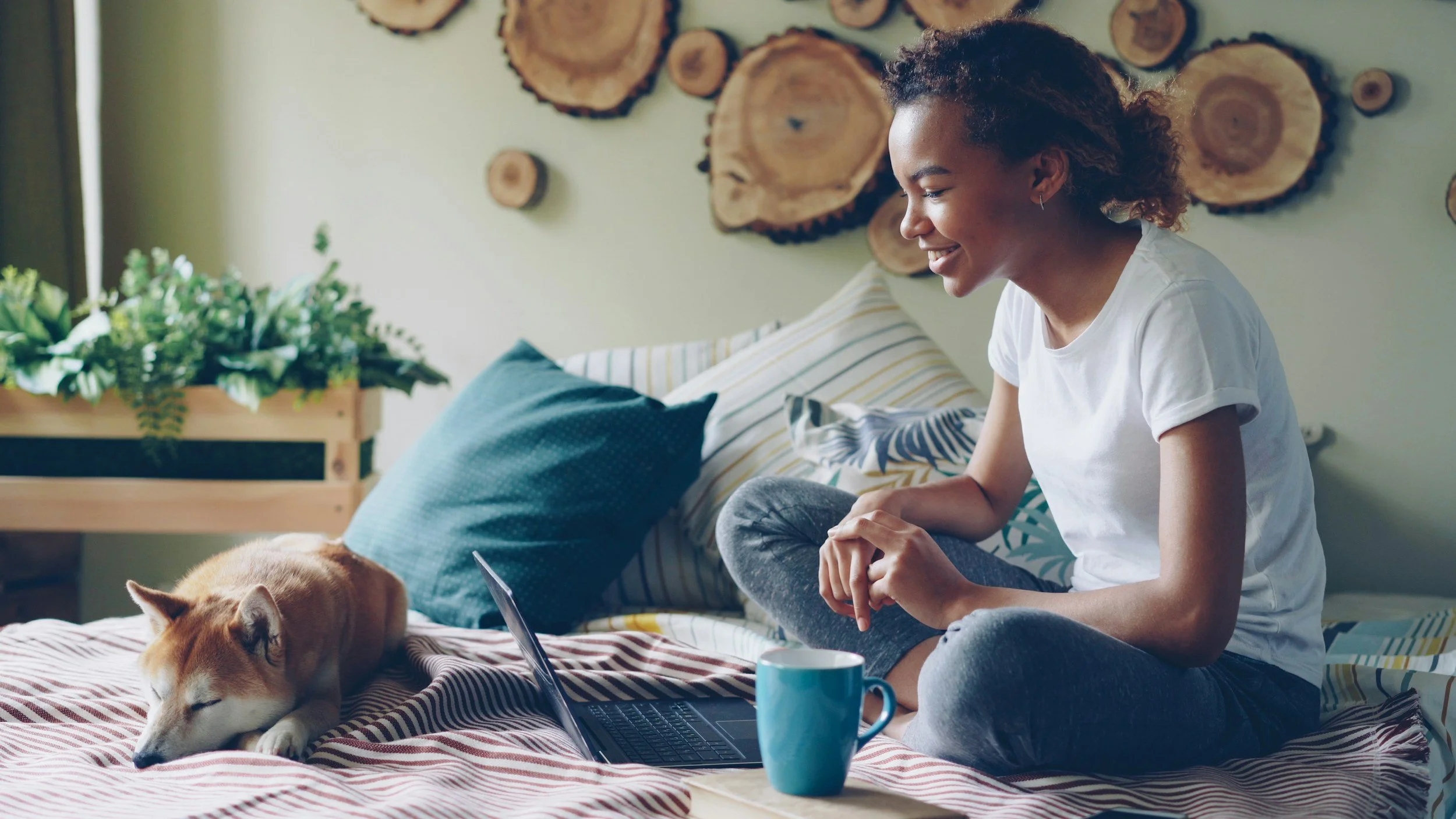 A woman sitting on her bed with a sleeping dog nearby, using a laptop, with a coffee mug in front of her and a decorative wooden wall hanging behind.