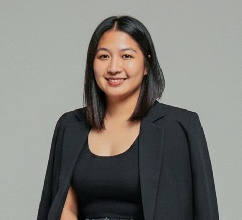 A woman with shoulder-length black hair, wearing a black top and a black blazer, smiling against a plain gray background.