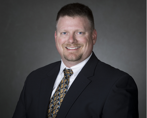 Professional portrait of a man in a dark suit, white shirt, and patterned tie, smiling against a gray background.