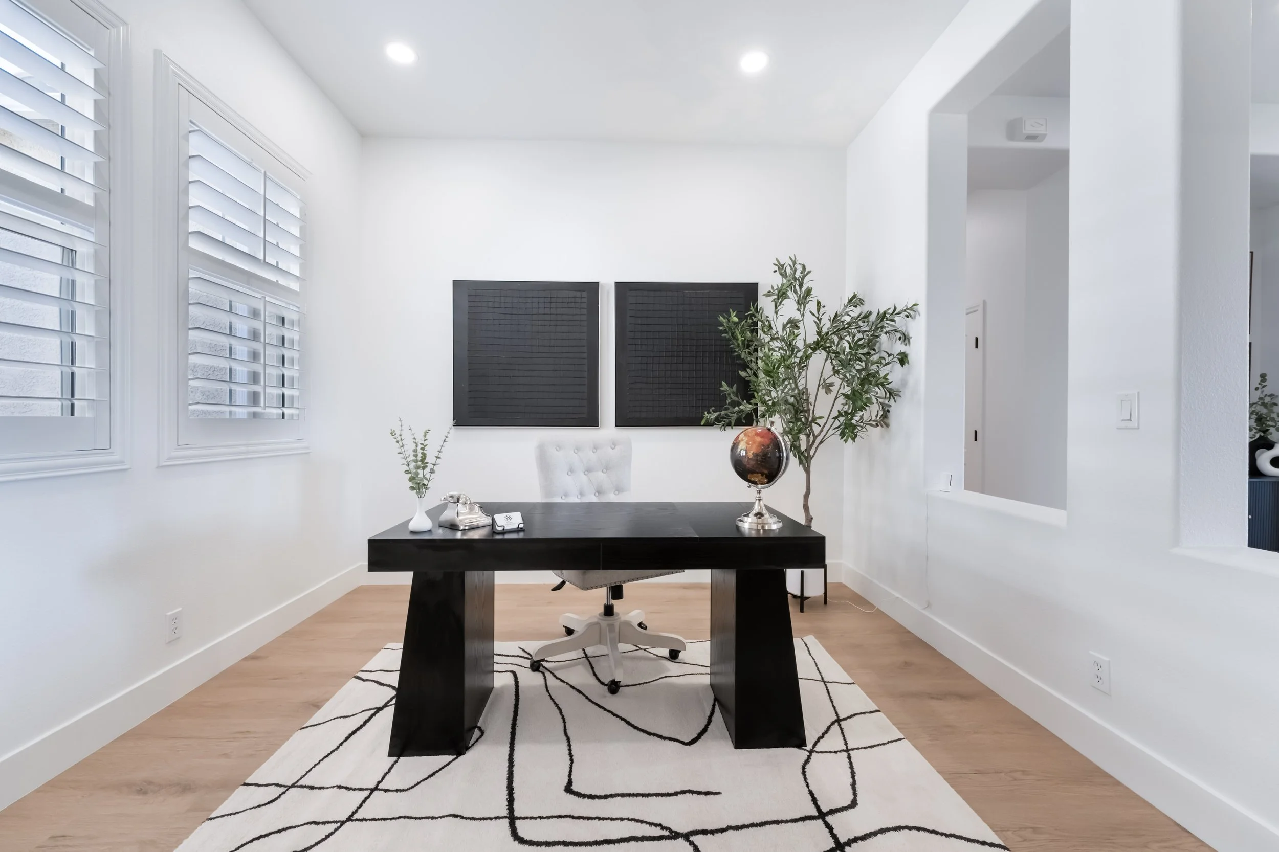 Modern office with white walls, black desk, white office chair, framed black squares on wall, large window with white blinds, potted plant, and patterned rug.
