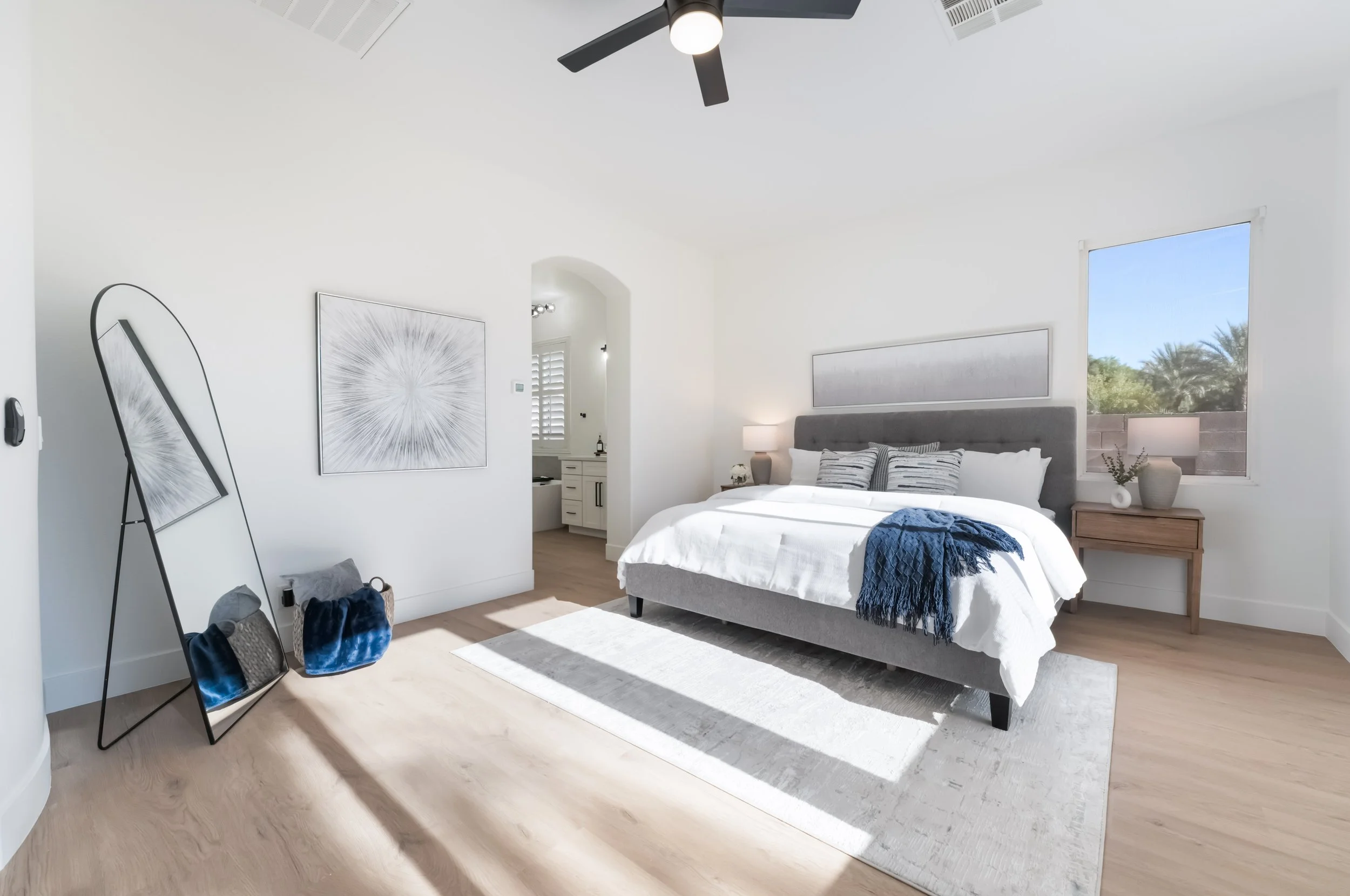 Modern bedroom with white walls, large bed with gray headboard, white bedding, blue throw blanket, and striped pillows. Two nightstands with lamps, a large window with a view of trees, and a ceiling fan. Decorative artwork on walls and a full-length mirror leaning against the wall.