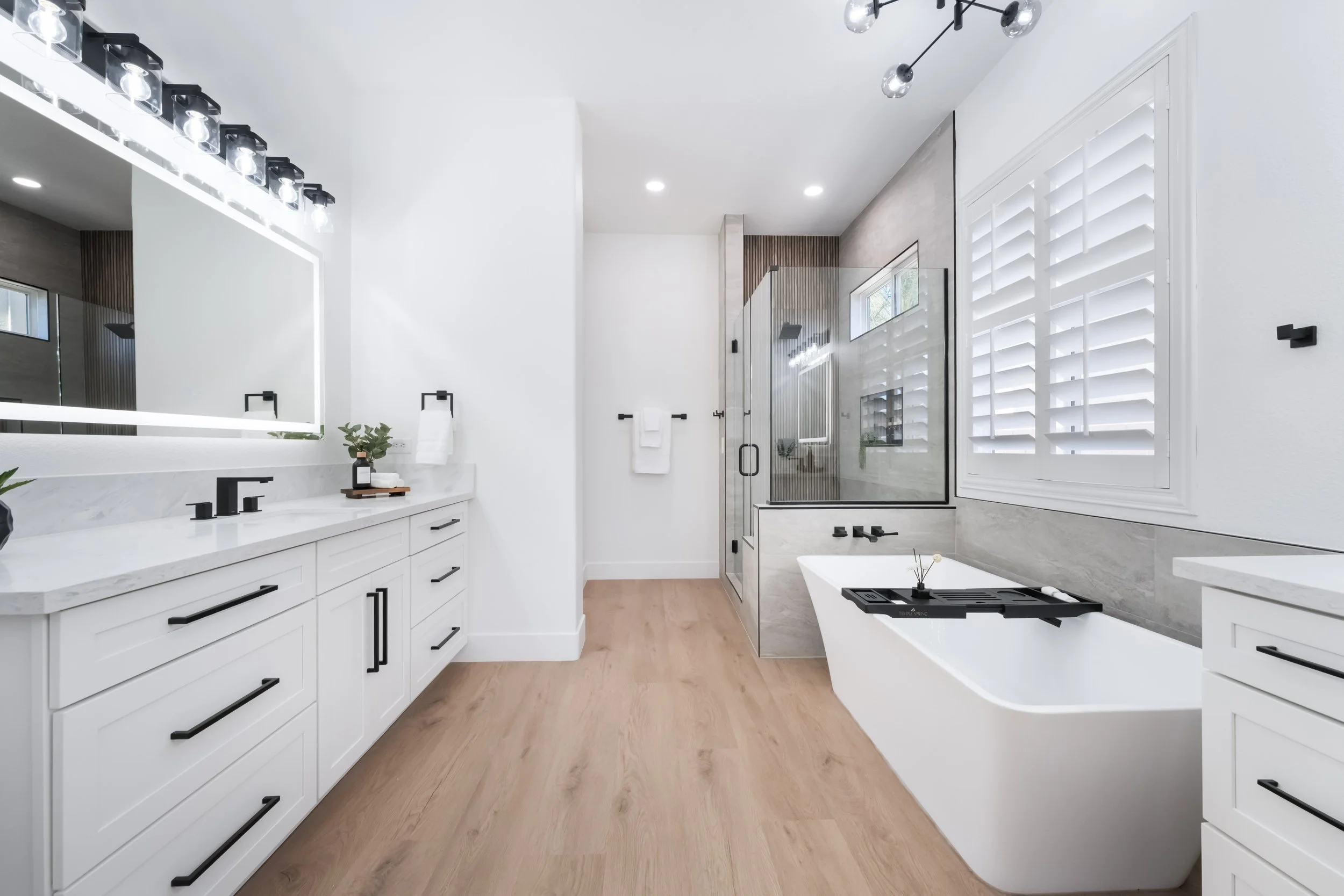 Modern bathroom with white cabinetry, a large mirror, wooden flooring, a bathtub with a black tray, and a glass-enclosed shower with wood accents.