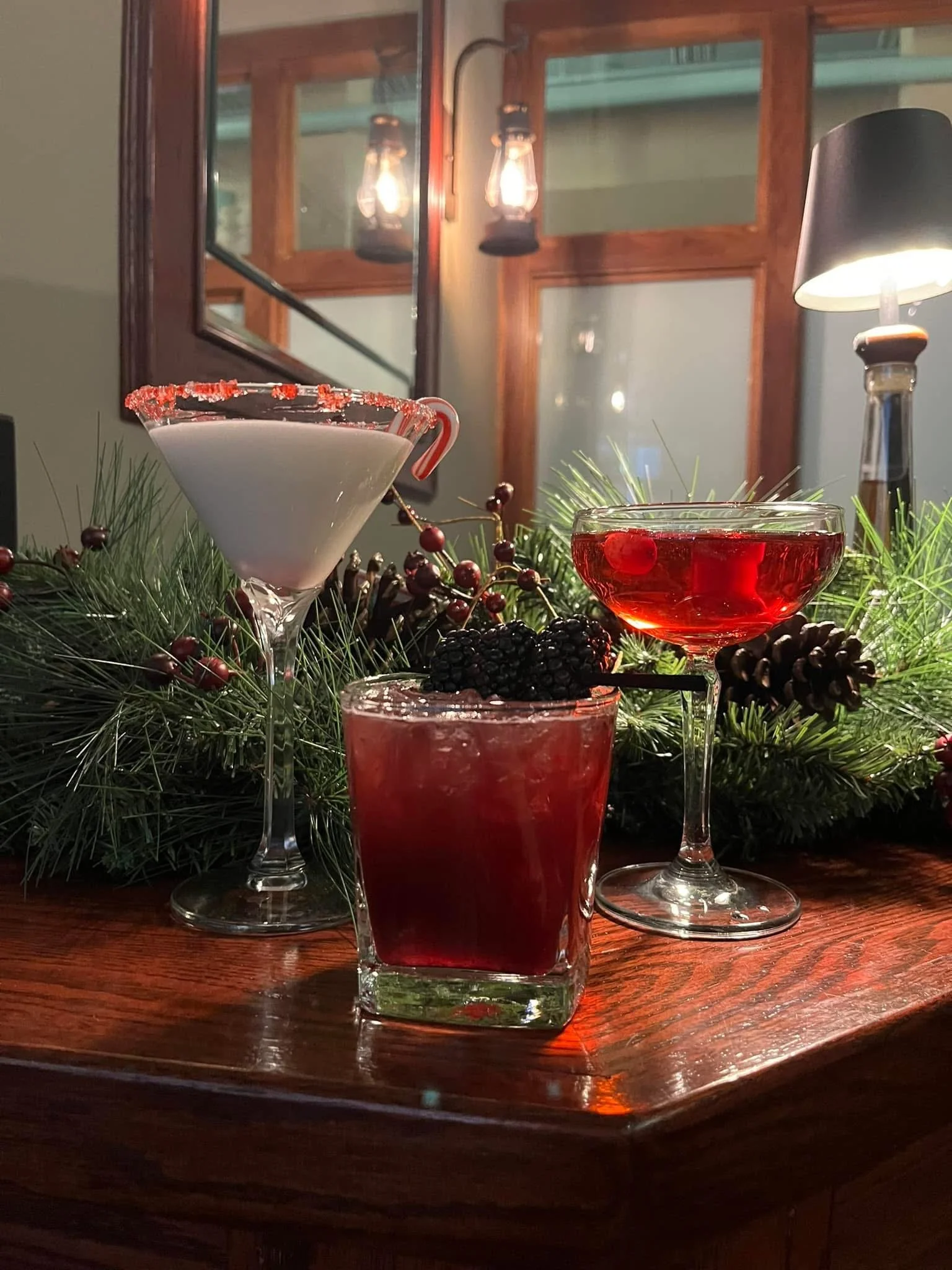 Three festive cocktails on a wooden table, decorated with pine branches and berries, with warm ambient lighting in the background.