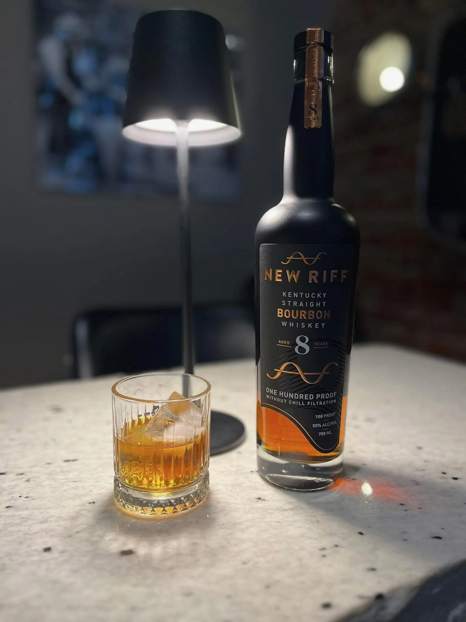 A bottle of New Riff Kentucky Straight Bourbon Whiskey and a glass with bourbon and ice on a white marble surface, with a blurred lamp in the background.
