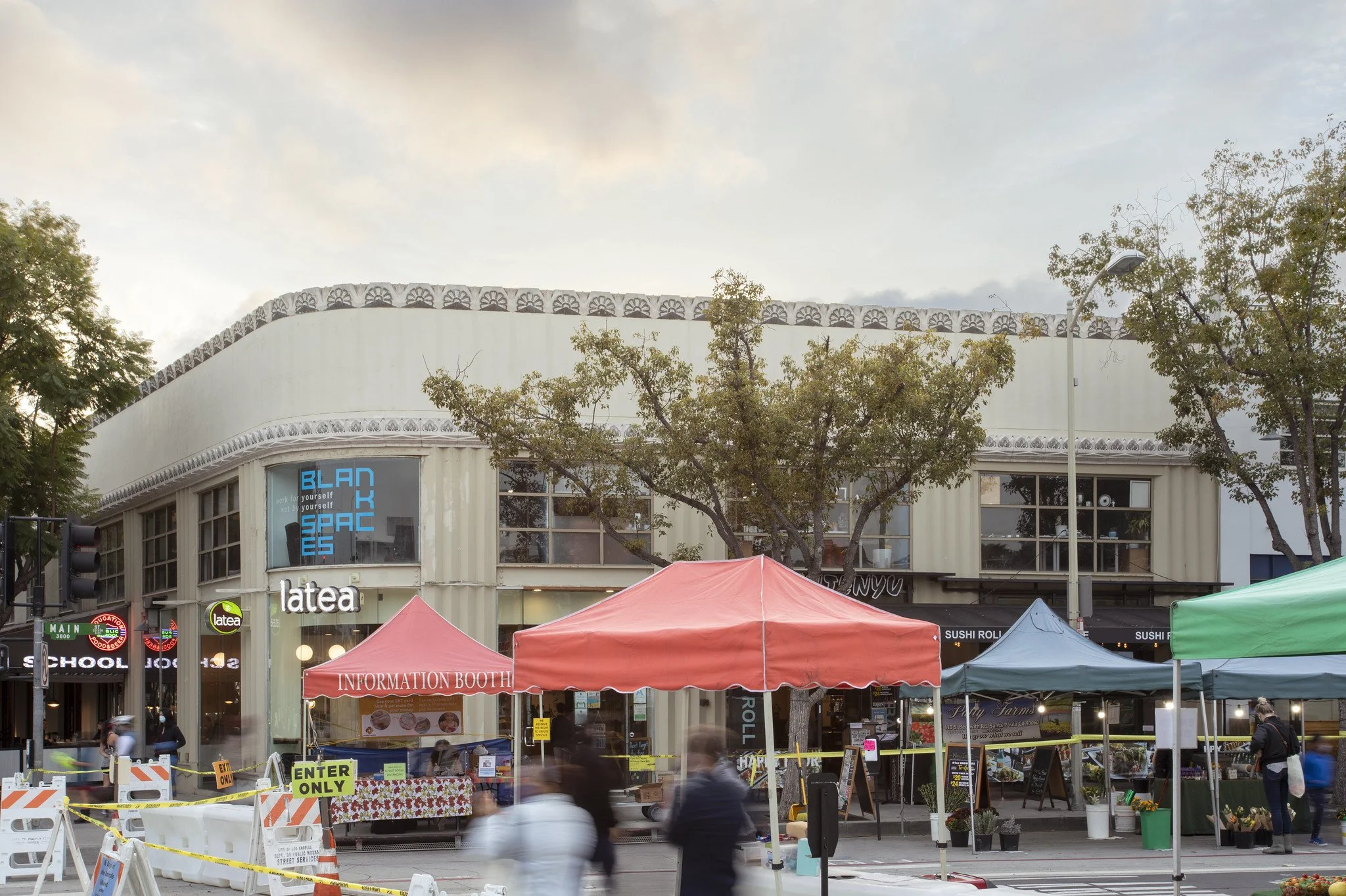 Outdoor street market with red, green, and blue tents, people walking, and a modern building with large windows and signs that say 'latea' and 'Blanc Spaces'.