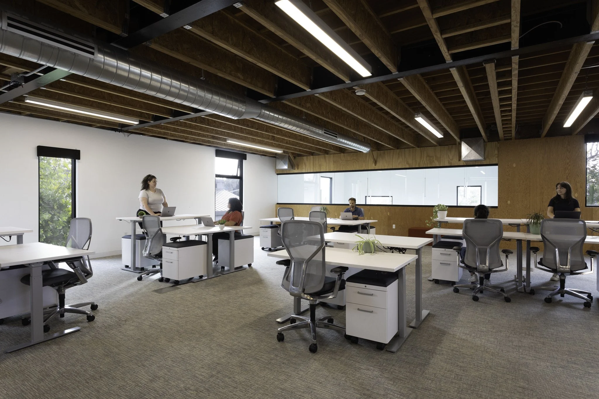 An office with several white desks and gray office chairs, some with plants, and four people working or conversing. The office has a high wooden ceiling with exposed beams and ductwork, large windows on one wall, and a long glass partition.