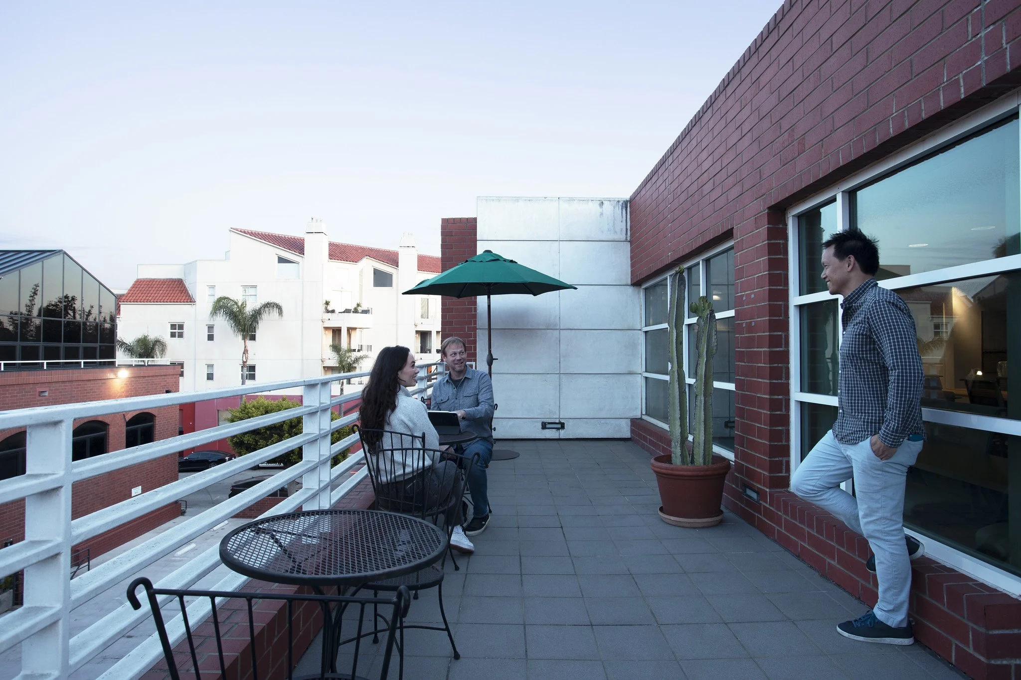 Three people on a rooftop terrace with a brick wall, green cactus, and an umbrella. Two are seated at a small table, one standing and leaning against the wall, engaged in conversation.
