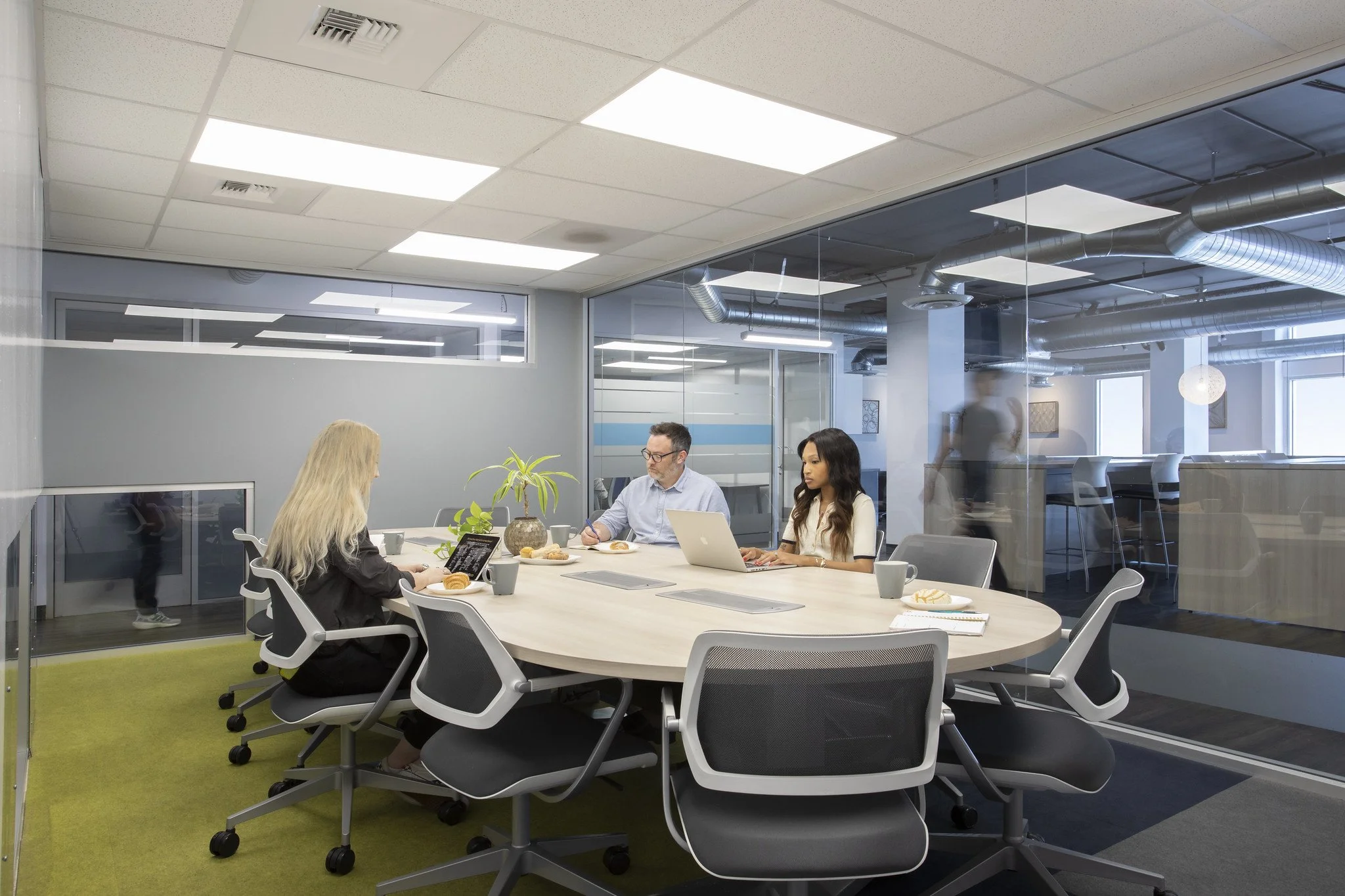 Business meeting in a modern conference room with three people seated at a large oval table, two women and one man, with laptops, notebooks, and coffee cups, behind glass walls with an open office space visible.