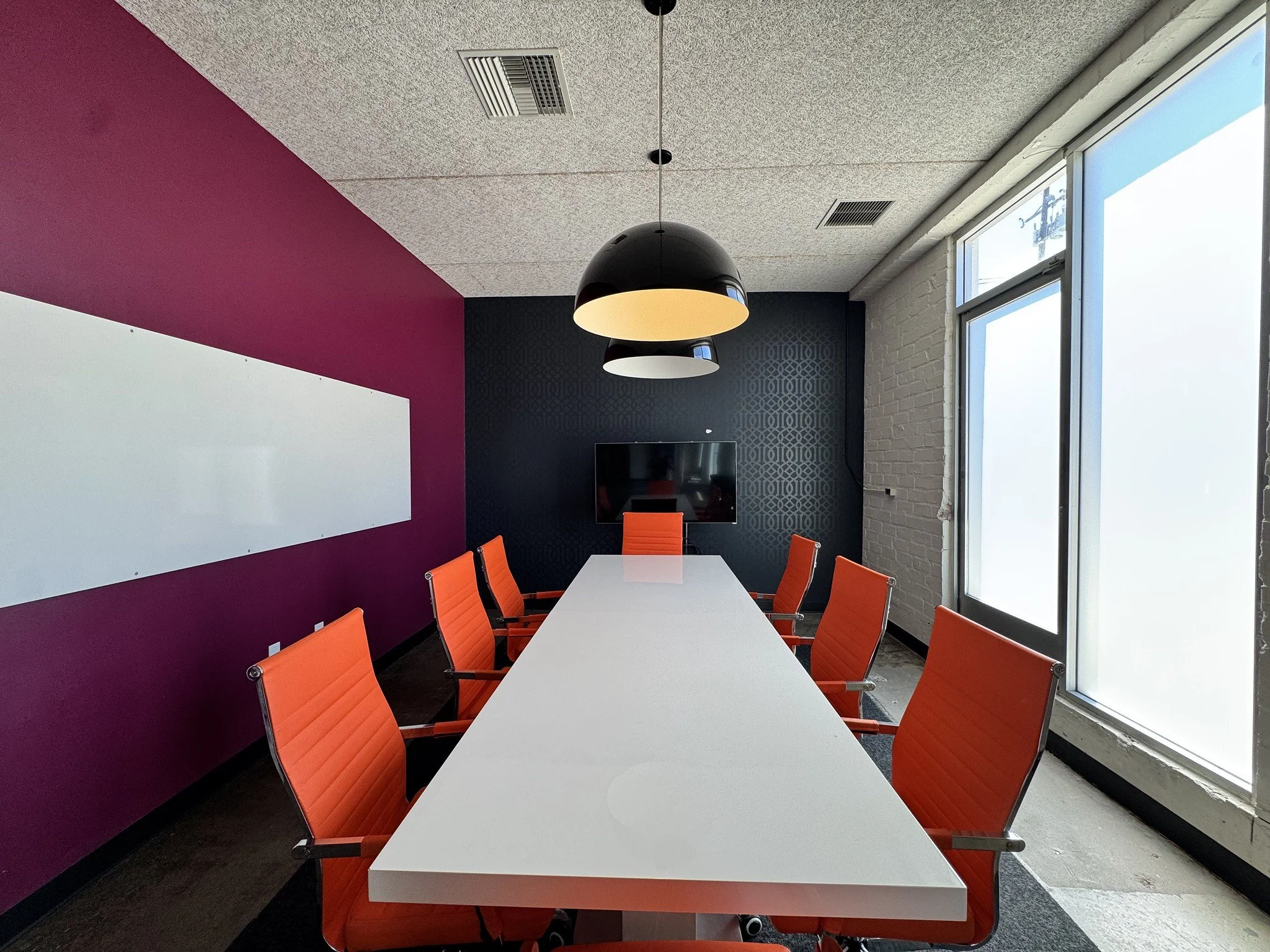 Modern conference room with a long white table, orange chairs, purple accent wall, black and white hanging pendant lights, a large window with frosted glass, a whiteboard, and a mounted television.