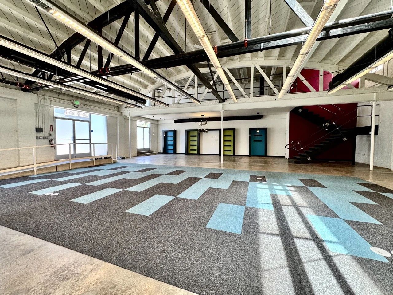 Empty interior room with blue and brown patterned carpet, white walls, high ceiling with exposed black steel beams and industrial lighting, glass door and windows on left, colorful doors and staircase on right.