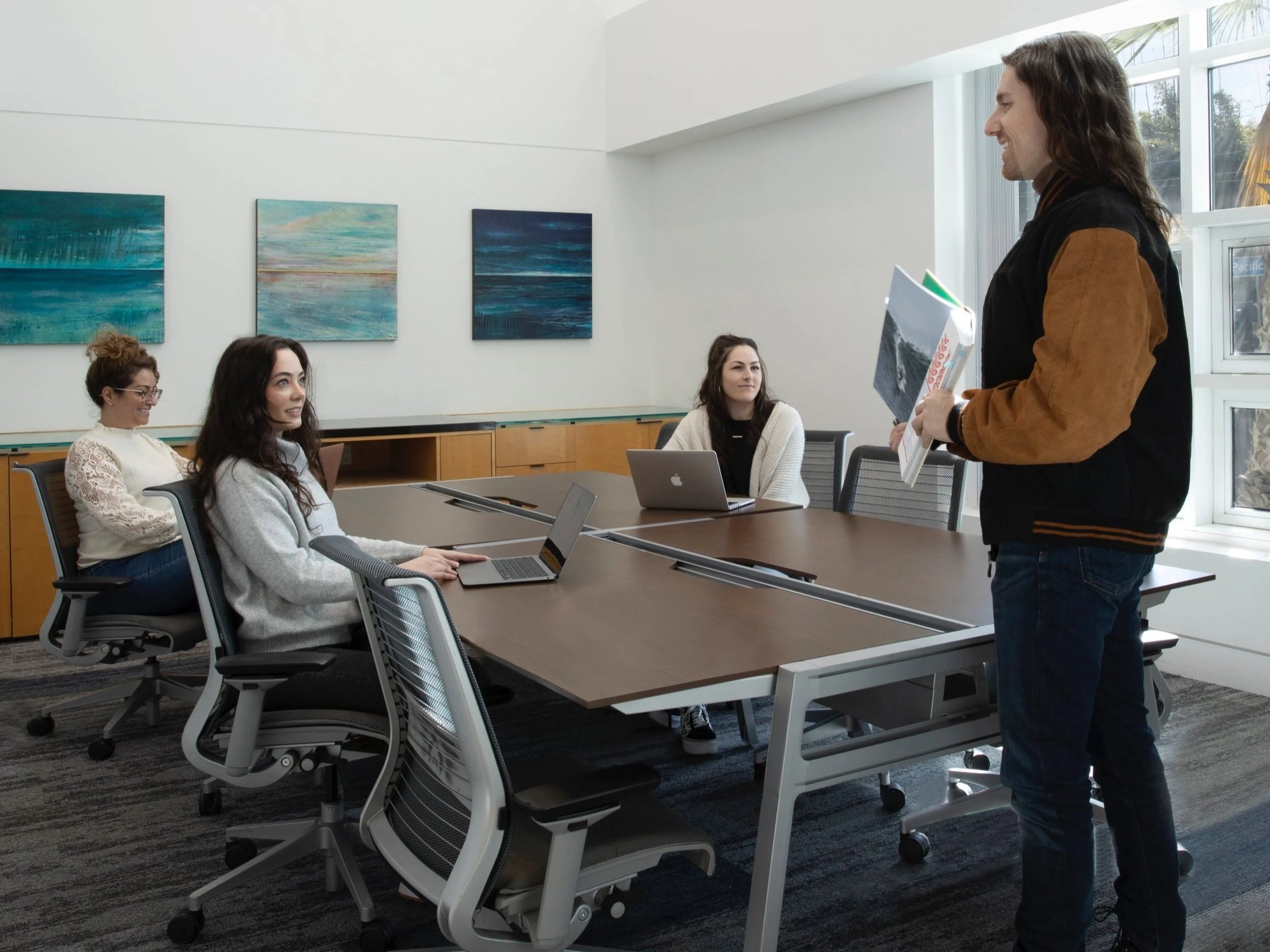 Four women sitting around a conference table with laptops, listening to a man standing and speaking in a meeting room.