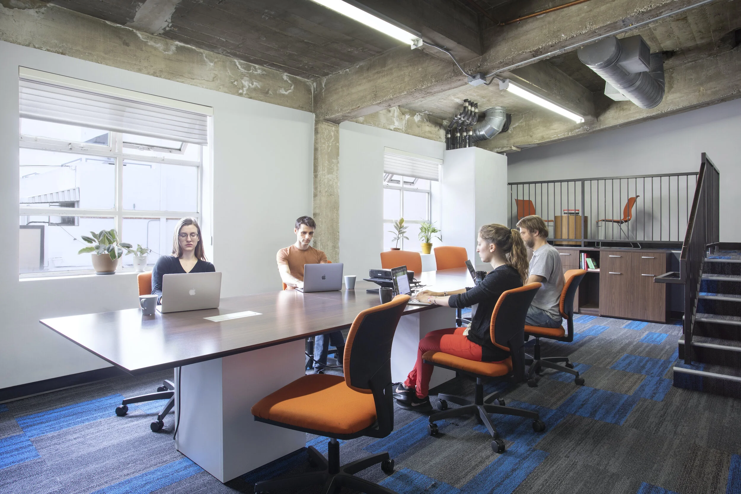 A modern office conference room at Blankspaces, CA, with five people working on laptops at a long table, large windows, potted plants, and industrial-style ceiling with exposed ductwork.