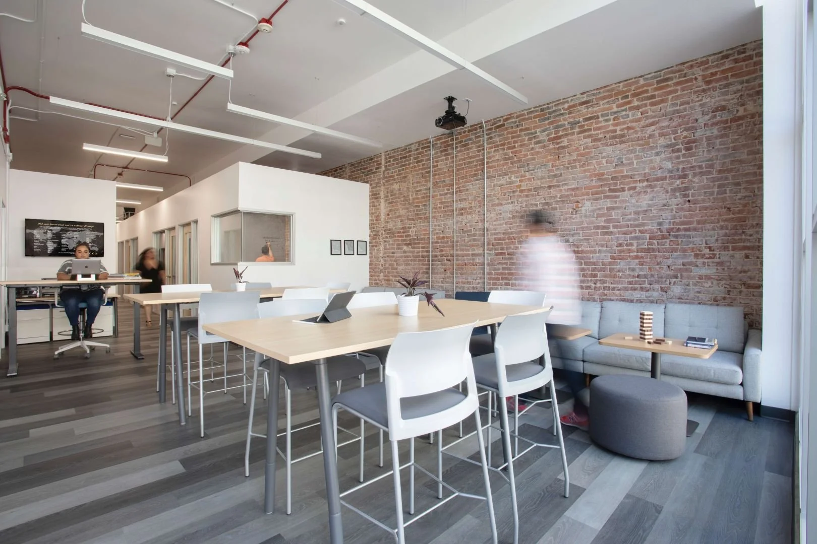 Modern office lounge with exposed brick wall, a large wooden table surrounded by white chairs, a gray sofa with a small wooden table and stack of books, and people working and walking in the background.