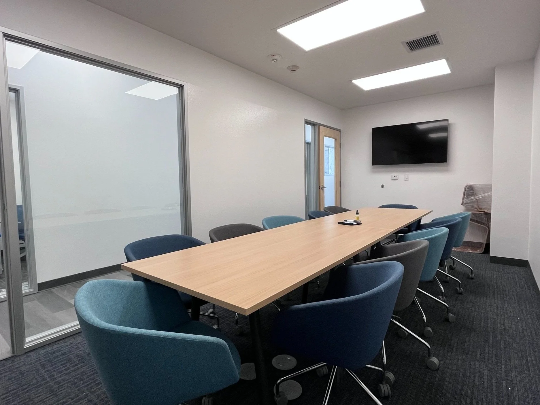 Empty conference room with a long wooden table surrounded by blue and gray upholstered chairs, a wall-mounted flat-screen TV, and a glass door leading outside.