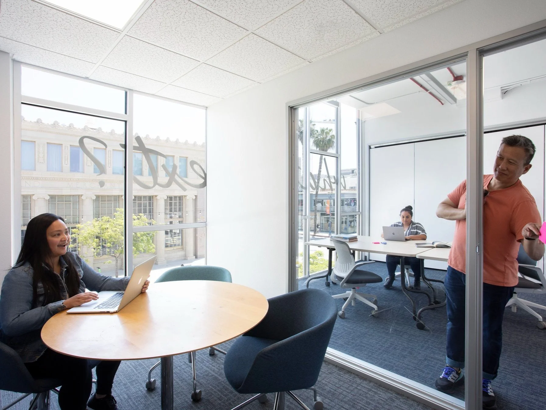 Two women working on laptops in a bright office, with a man writing on a glass wall, visible through a glass wall separating the rooms.