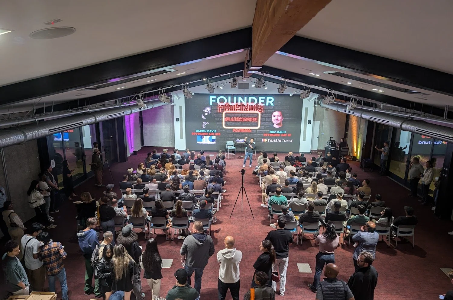 A large indoor conference room filled with people seated and standing, attending a presentation on stage. The stage has a big screen displaying the words "FOUNDER FRIENDS," photos of two speakers, and additional text, with colored lighting around the room.