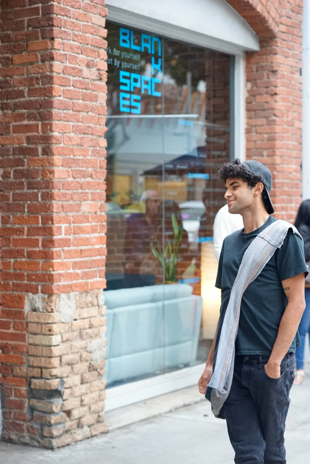 A young man stands on a city sidewalk smiling and looking at a storefront with a neon sign that reads 'BLACK SPACES'. The storefront has large glass windows and a red brick exterior.