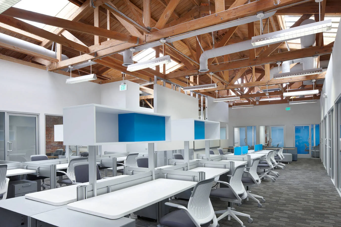 Empty modern office space with desks and chairs, exposed wooden ceiling beams, and large windows.