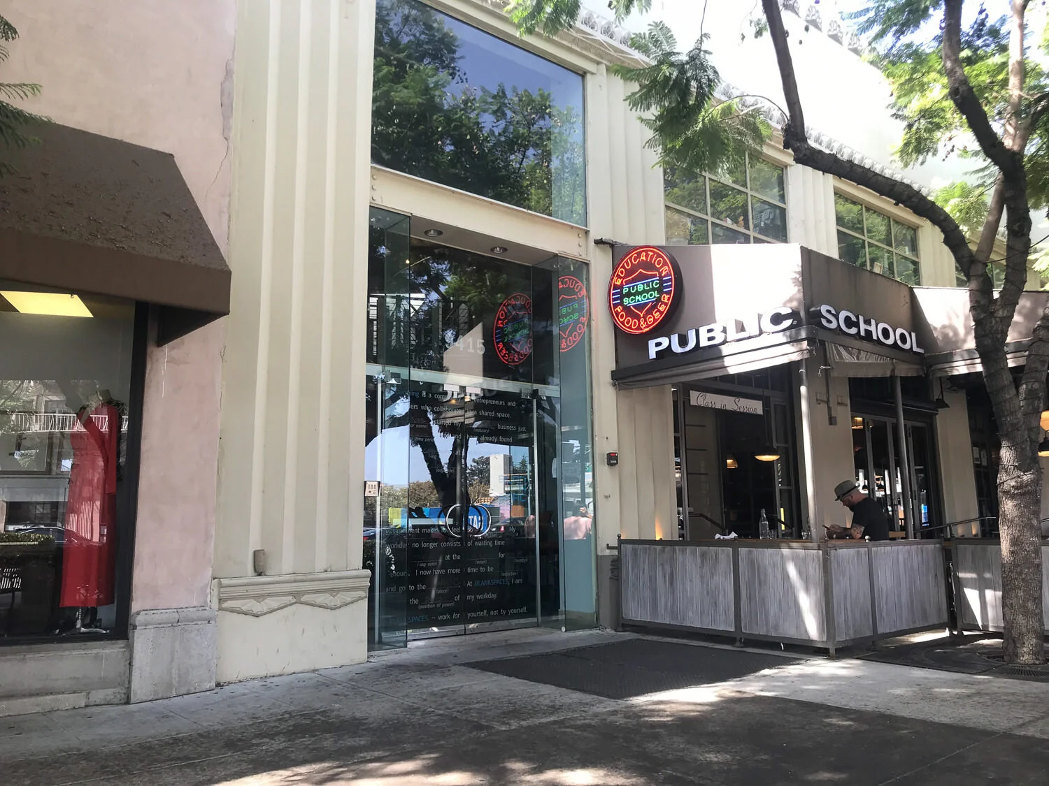 Street view of a building with a sign that reads 'Public School' and glass windows reflecting trees, with a person sitting outside on a patio using a phone, and trees lining the sidewalk.