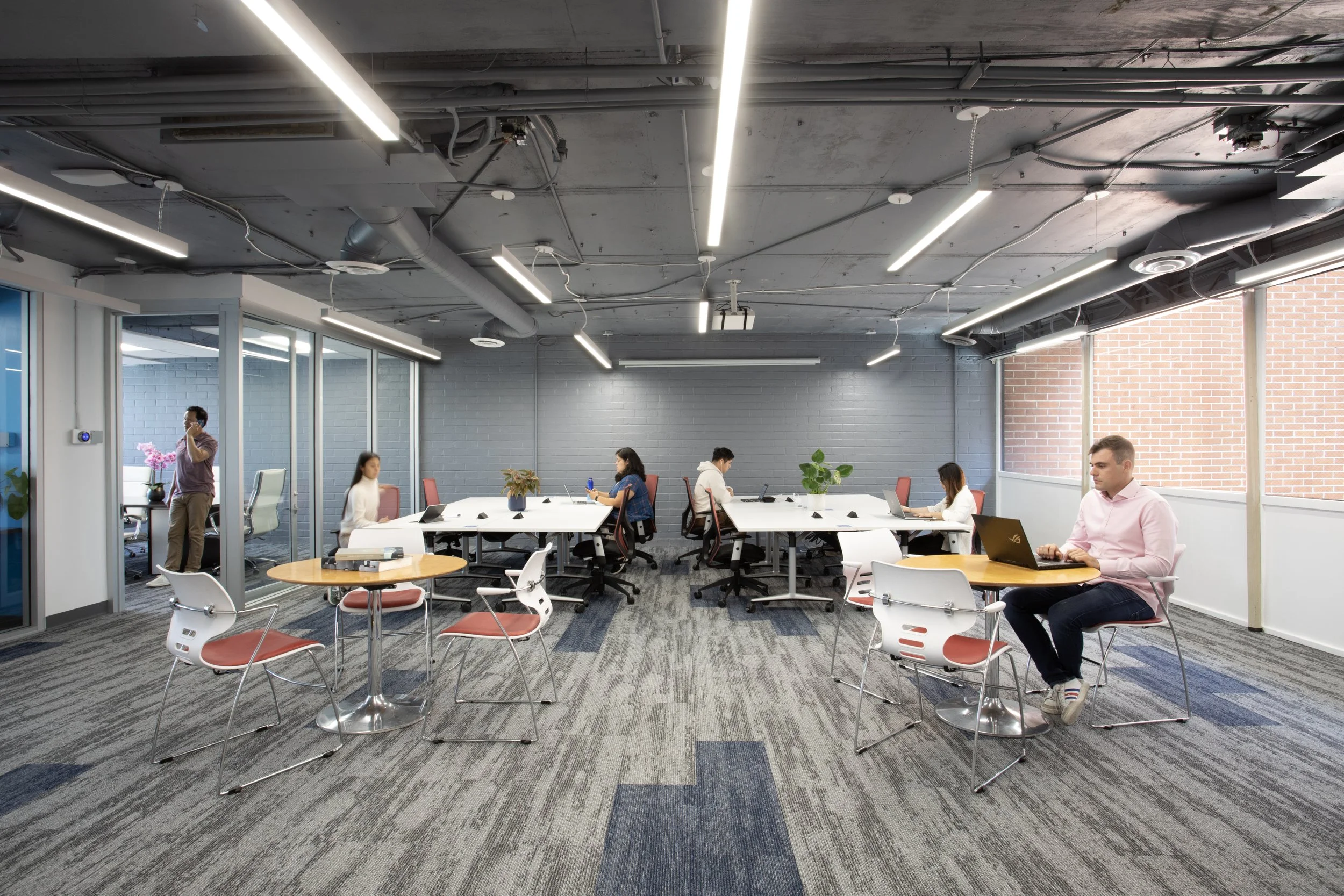 Blankspaces office space with multiple employees working at desks and a man sitting at a small round table using a laptop. Glass walls, gray walls, exposed ceiling with lighting fixtures, and large windows with brick exterior visible.