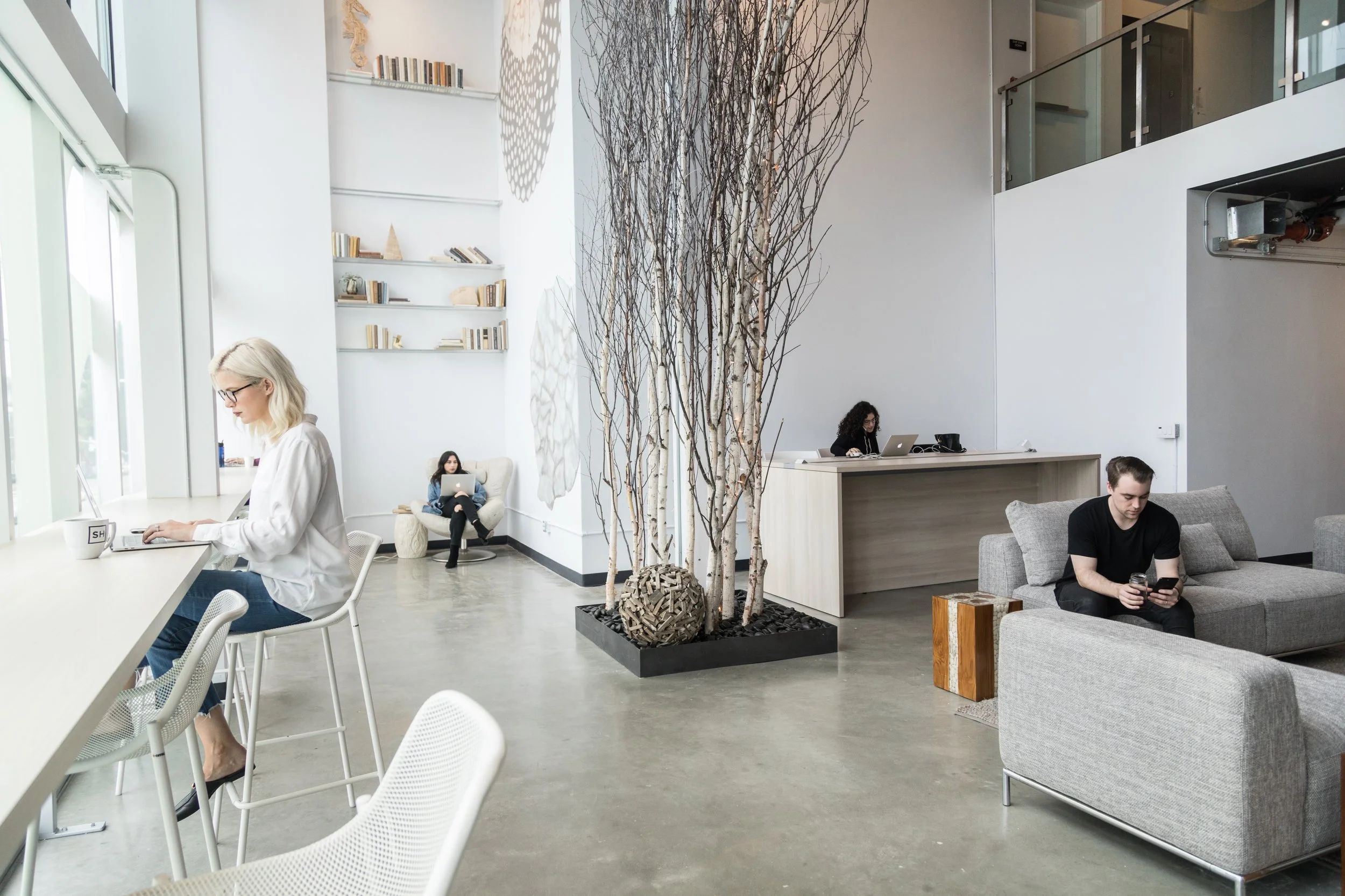 Modern lounge area with white walls and large windows, featuring a woman working on a laptop at a high table, a woman sitting on a white armchair using a laptop, a man sitting on a gray sofa looking at his phone, and a woman working at a desk behind 