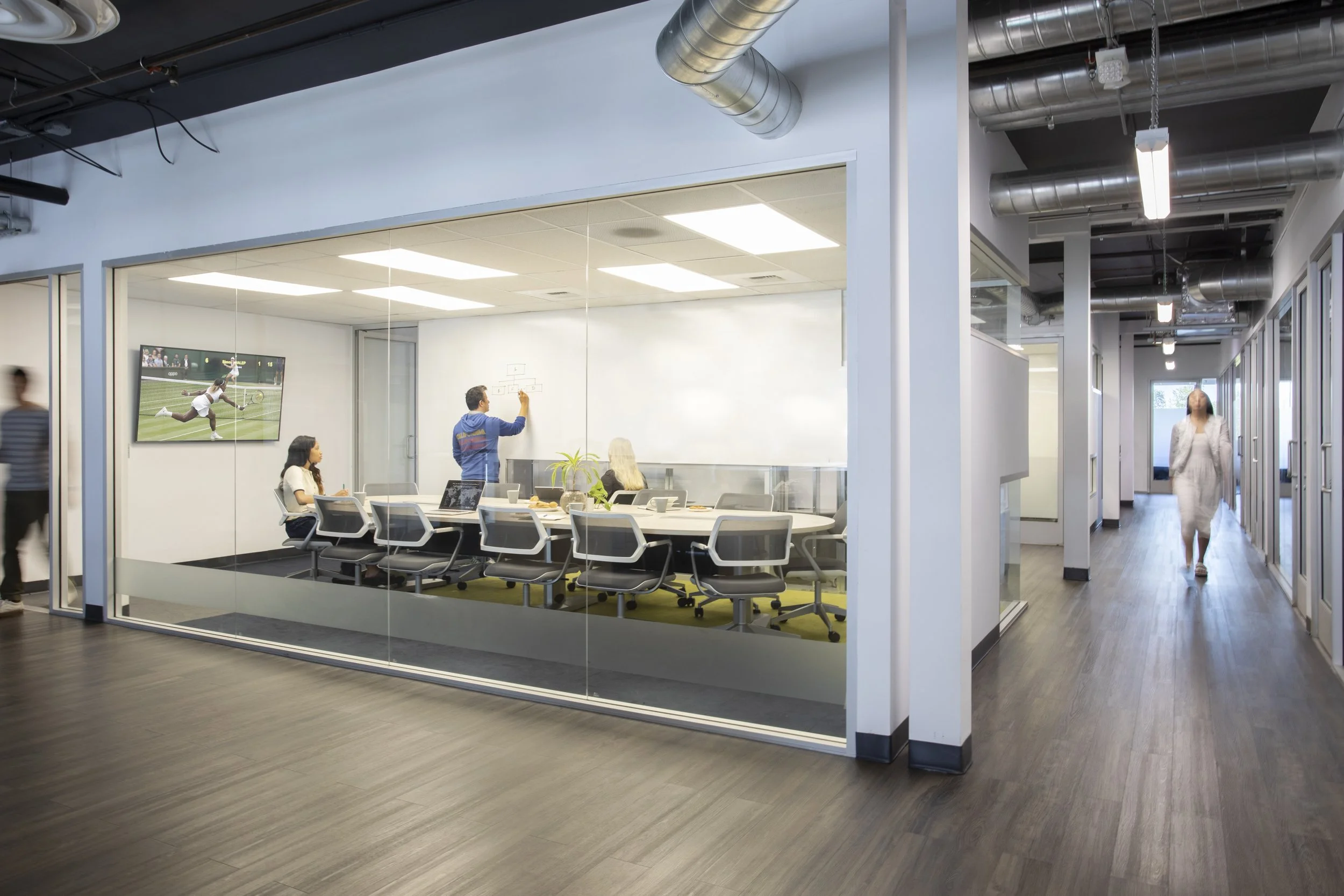 Office conference room with a man drawing tactics on a whiteboard, three women seated around the table, a large TV screen showing a tennis match, and people walking down the corridor outside.