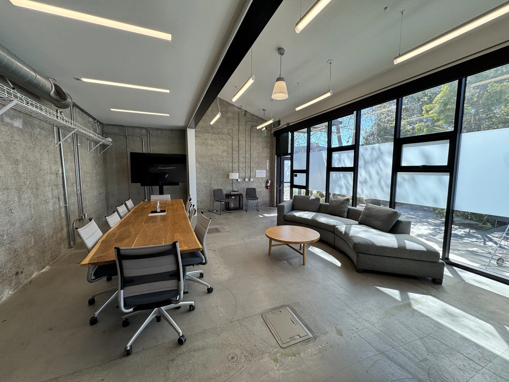 Modern office conference room with a large wooden table, ergonomic chairs, a TV screen, a gray curved sofa, a round coffee table, and large windows.