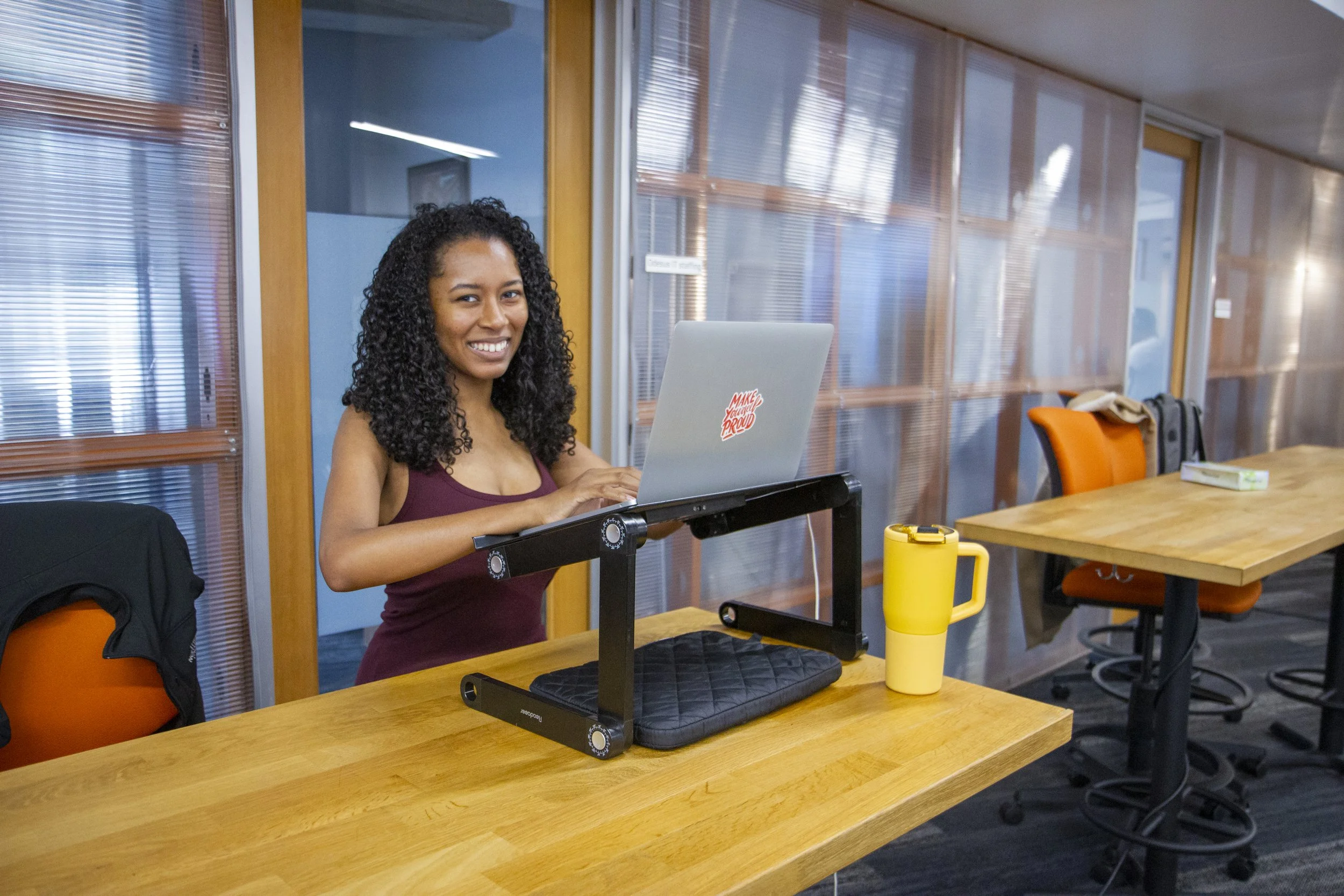 A woman with curly black hair working on a laptop in a conference room with orange chairs and wooden tables.