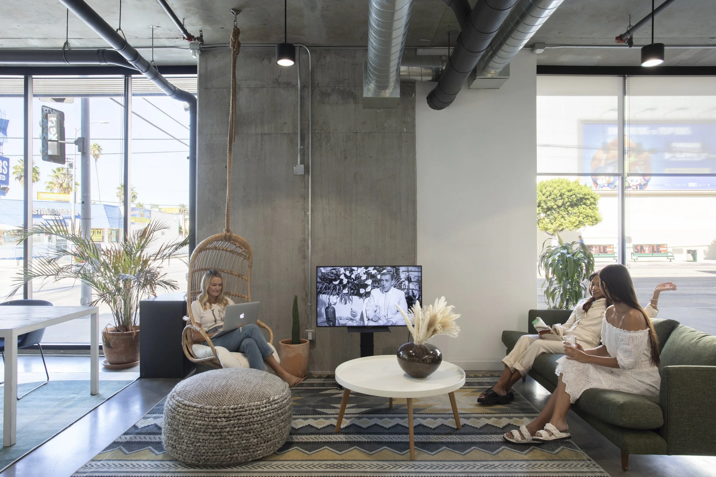 Three women in a modern cafe, one sitting on an armchair using a laptop, one on a sofa with a phone, and the third holding a drink, with a TV on a stand, plants, and large windows in the background.