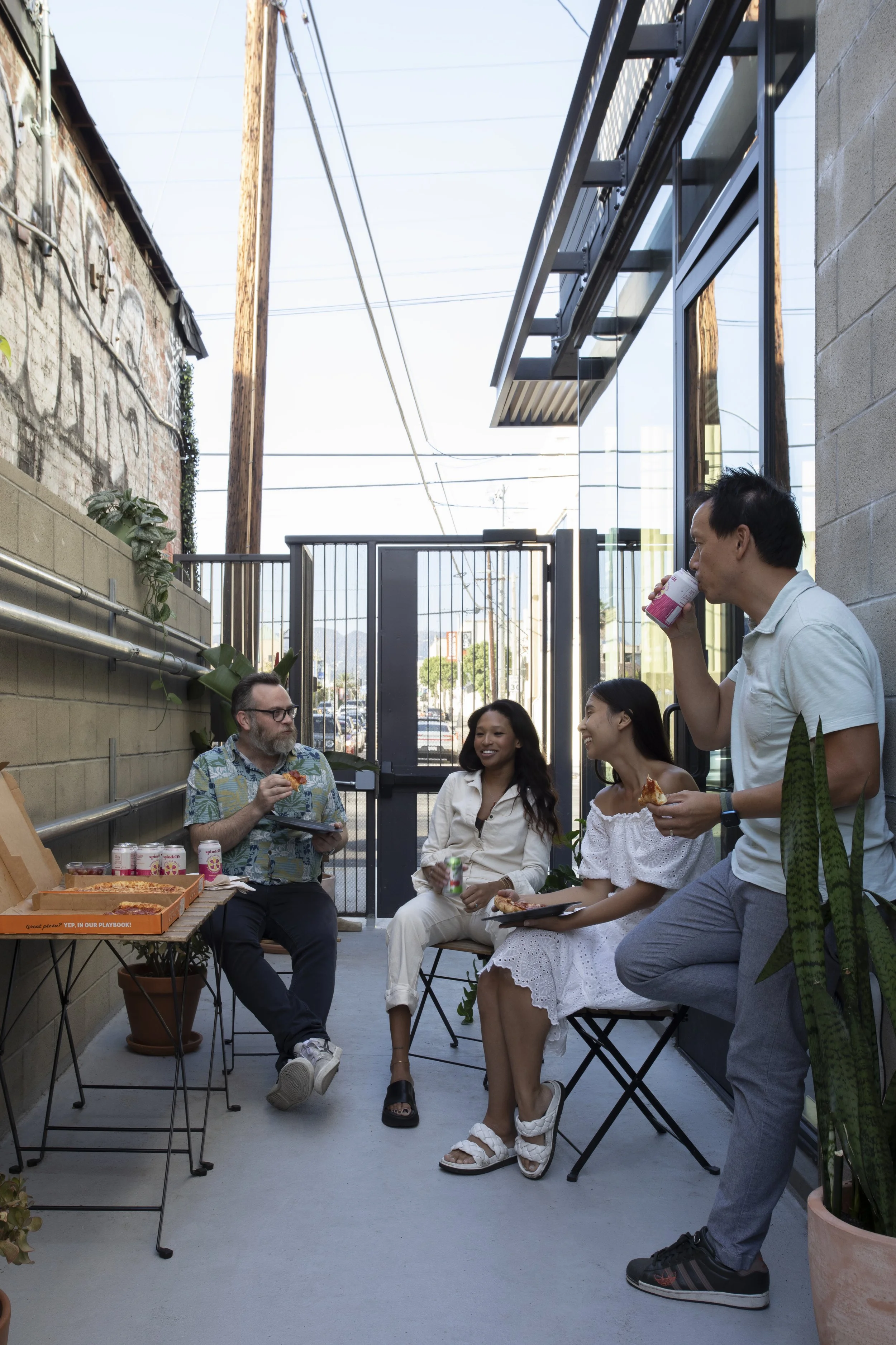 Four people enjoying pizza outdoors on a patio, with three sitting and one standing, while drinking from cans.