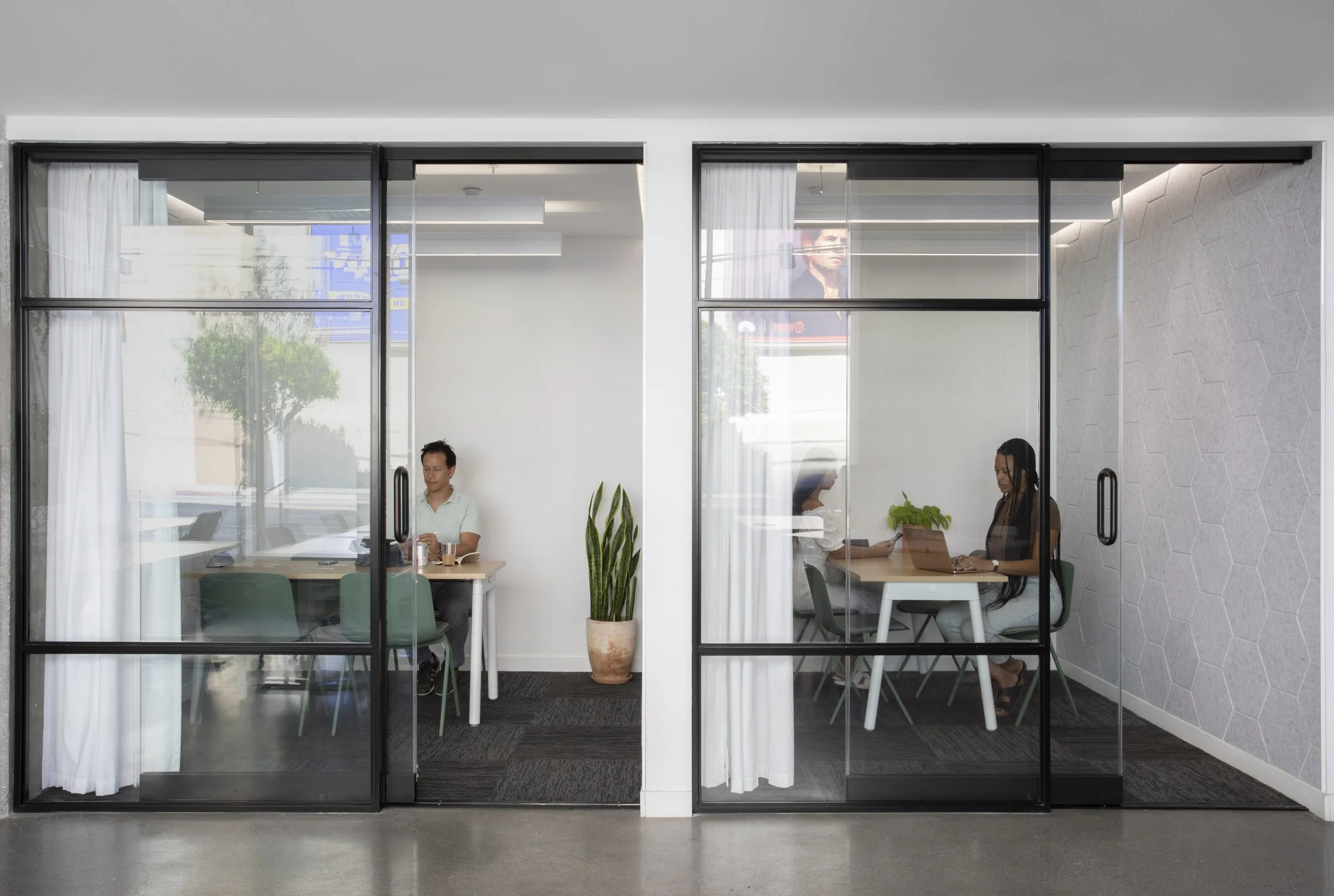 Two separate enclosed office spaces separated by glass walls and doors, each with individuals working at tables with laptops. The left office has a man sitting alone, and the right office has two women working together. There are potted plants, white curtains, and minimalist decor inside.