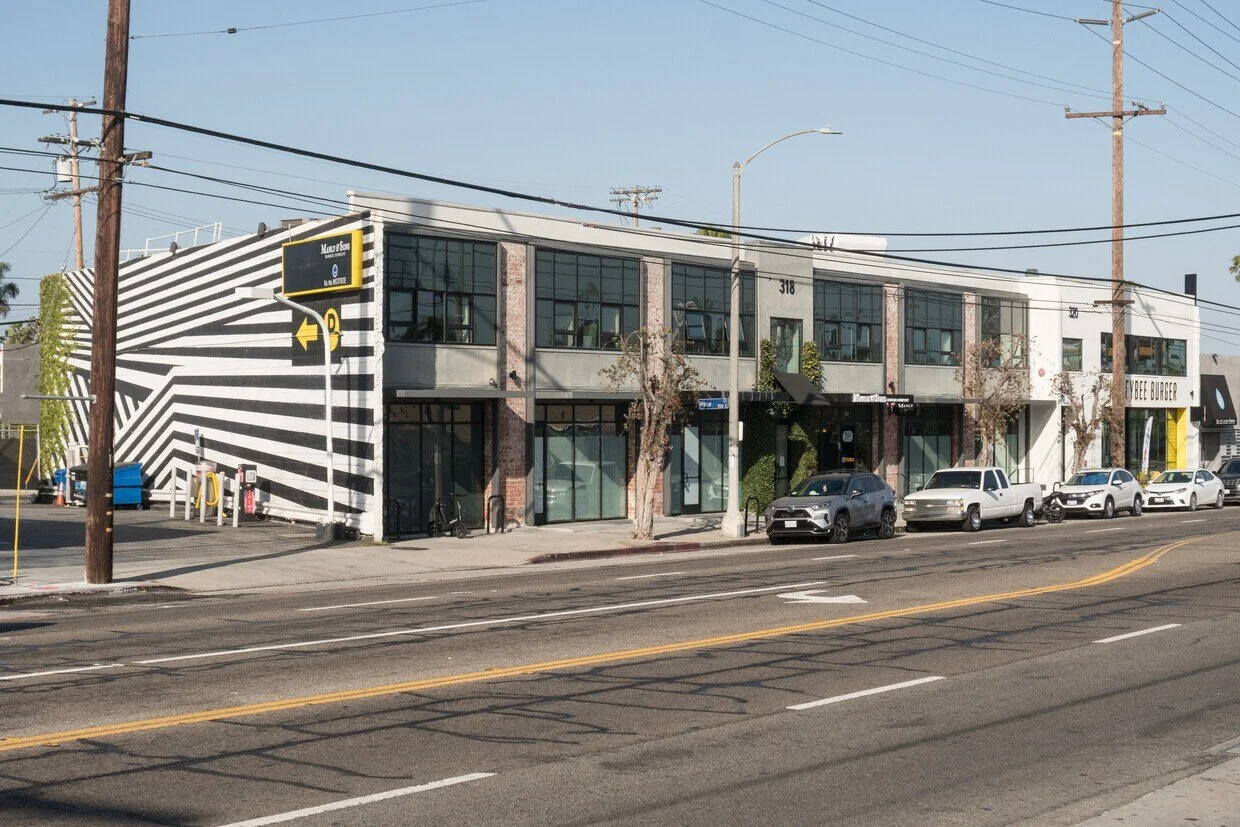 Street view of a Blankspaces office building, featuring large windows and a distinctive black and white striped mural on one side. Several cars are parked along the sidewalk, and utility poles with power lines line the street.