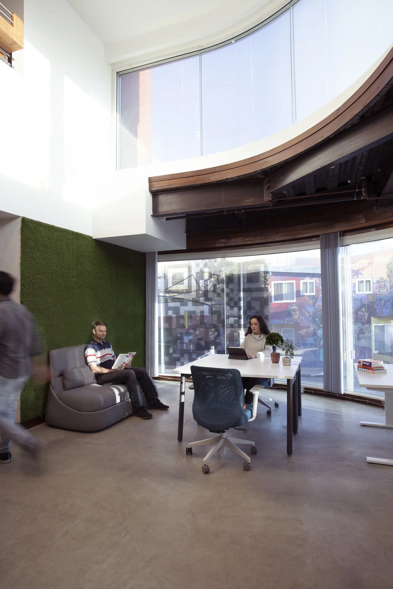 Modern office space with large windows, a green wall, and a mix of seating including a sofa and a desk with chairs. Two people are visible, one seated at the desk working on a laptop and another sitting on the sofa reading a magazine.
