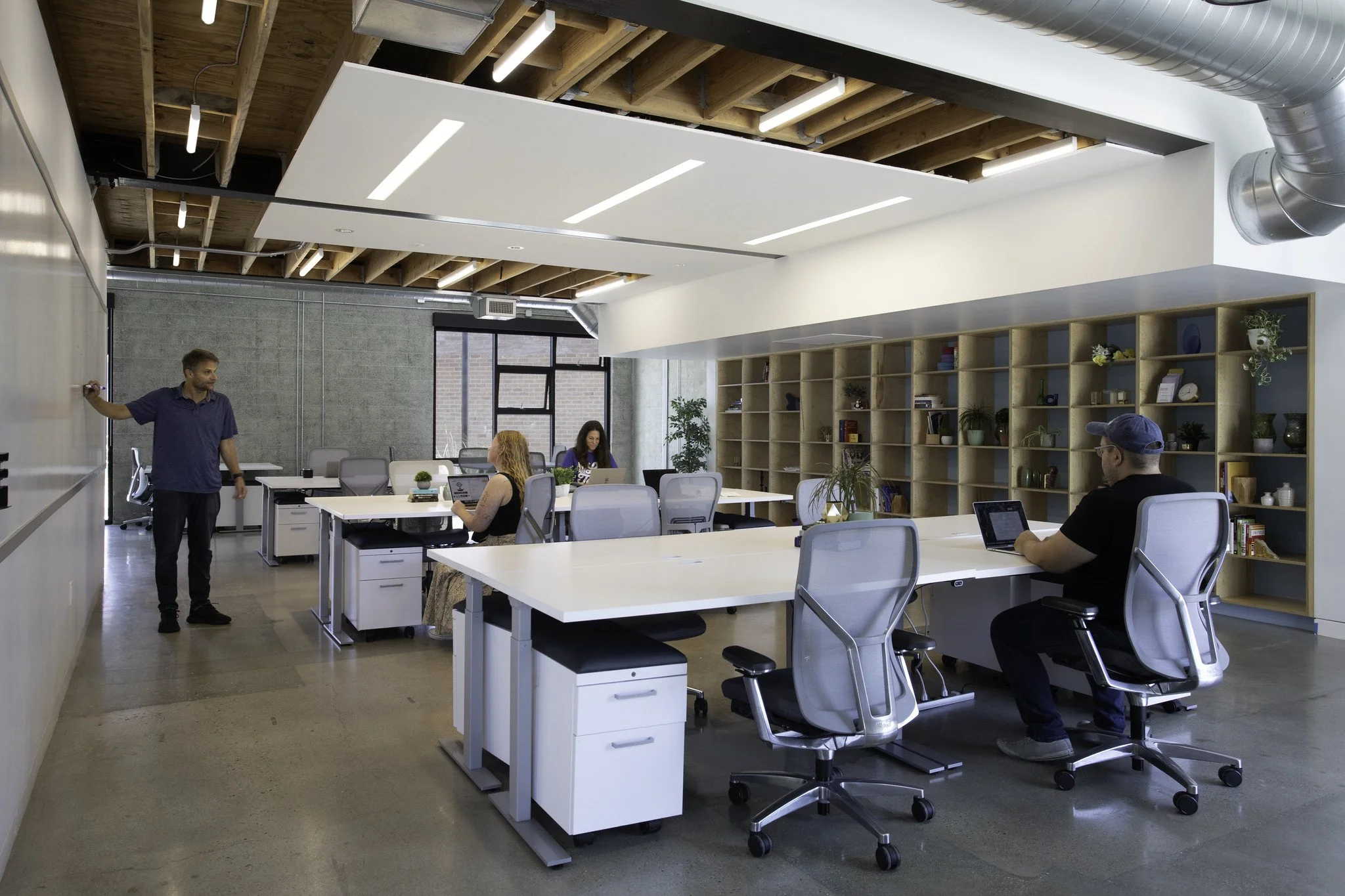 Modern open-plan office with exposed wooden ceiling beams, white desks, gray ergonomic chairs, and a large bookshelf filled with decorative items and plants. Four people are working, one standing and three seated, with large windows providing natural