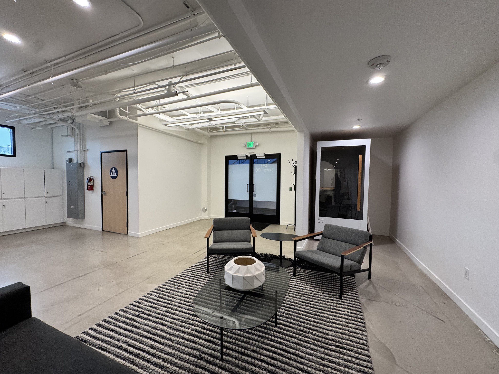 Modern office lounge area with two gray armchairs, a black coffee table with a decorative white vase, a striped rug, and a black-framed glass display case. In the background, there are double glass doors, a fire extinguisher, lockers, and a restroom 