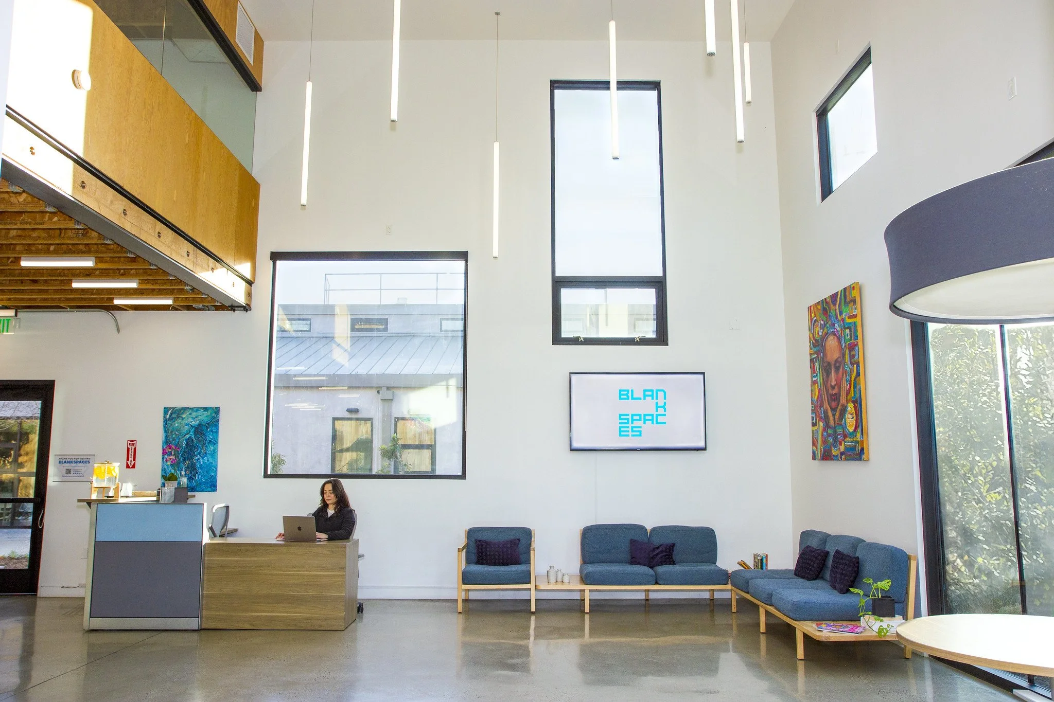 Modern lobby area with a woman sitting at a reception desk, blue couches with purple pillows, large windows, colorful artwork, and a digital sign reading 'BLAD SPAC ES'.