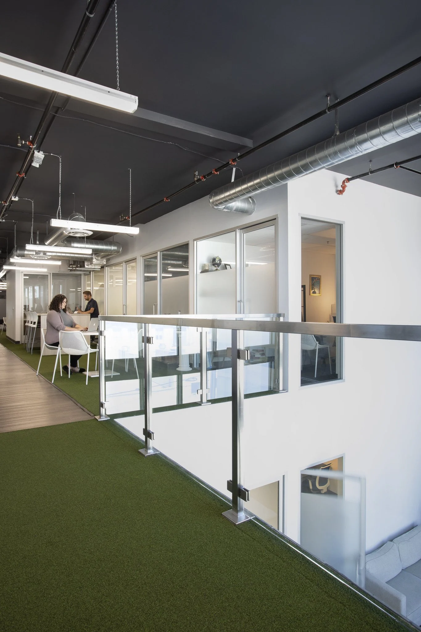 Modern office space with glass partitions, a woman working at a desk, and another person standing nearby, with industrial-style ceiling and green carpet flooring.