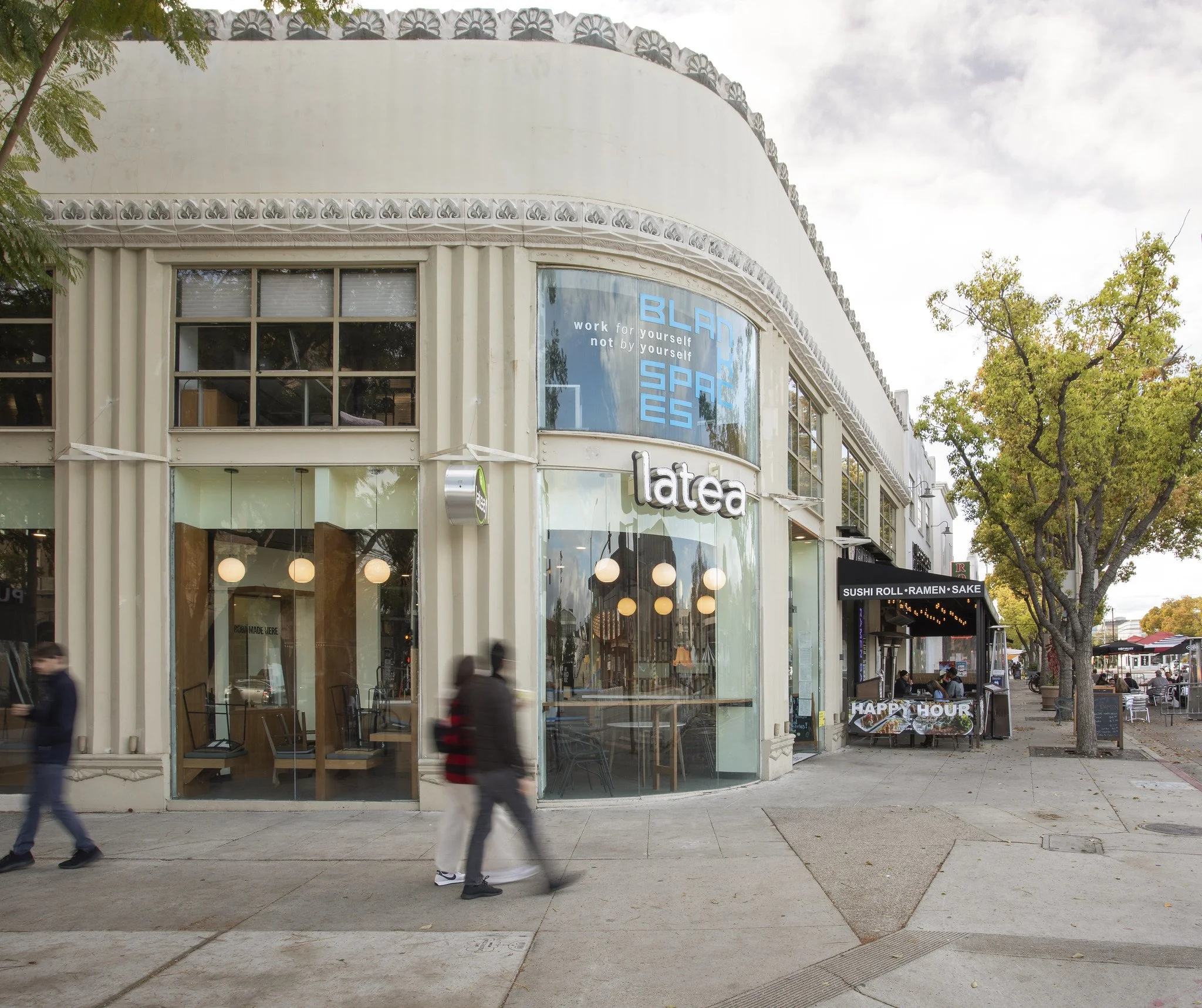 Street view of a cafe named 'LATEA' with large glass windows, patio seating, and trees lining the sidewalk. A man and woman are walking past, with a neighboring restaurant advertising sushi, ramen, and sake.