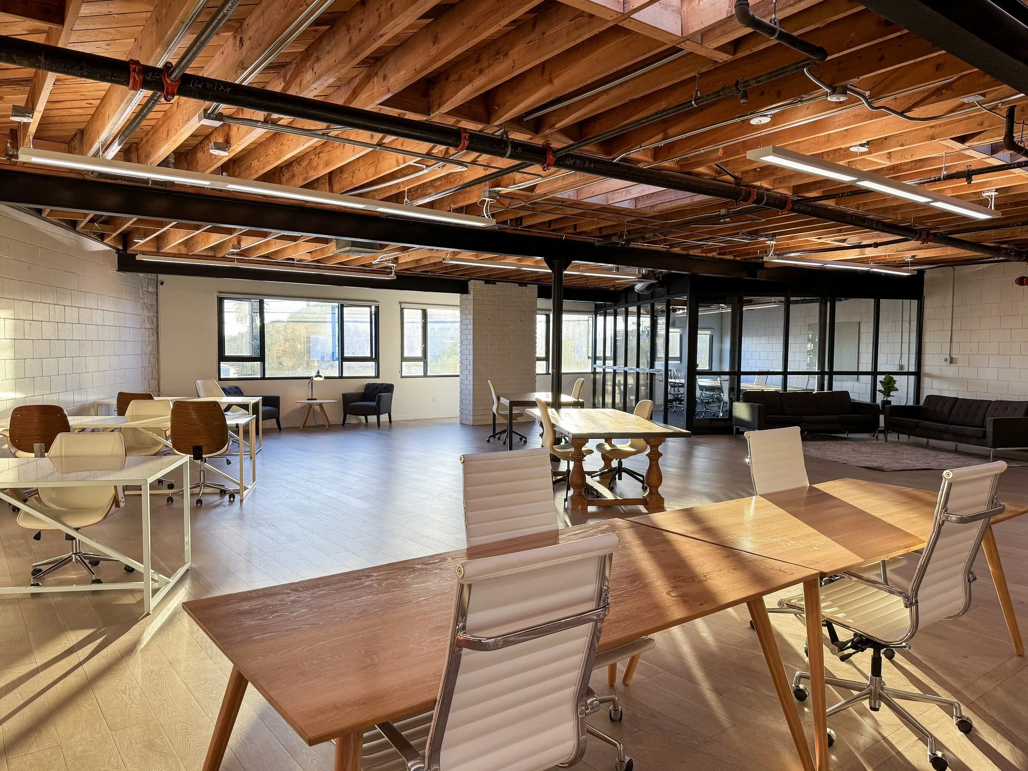 An open-plan modern coworking space with wooden ceilings, large windows, and various seating arrangements, including tables, chairs, and lounge areas.