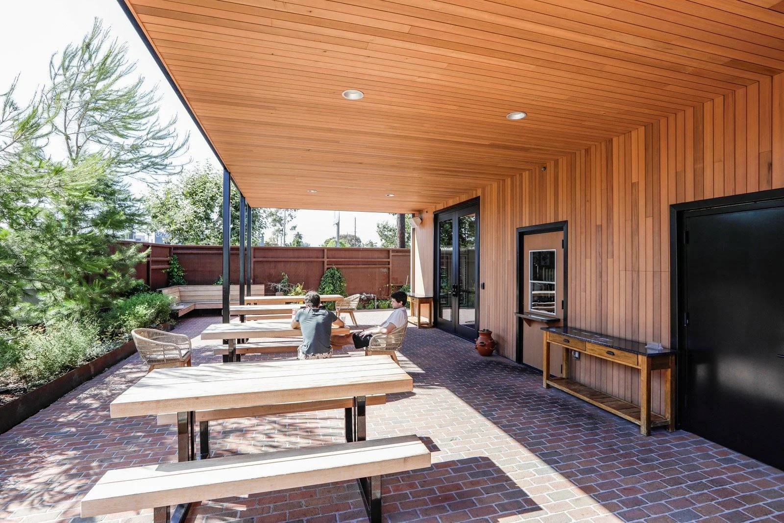Two people sitting at a picnic table on a covered outdoor patio with brick flooring, wooden walls, and ceiling, surrounded by trees and a red fence.