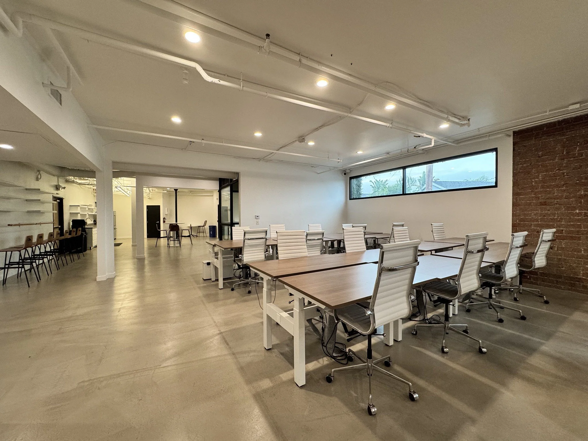 Modern conference room with white chairs and wooden tables, a long rectangular window, brick accent wall, and polished concrete floor.