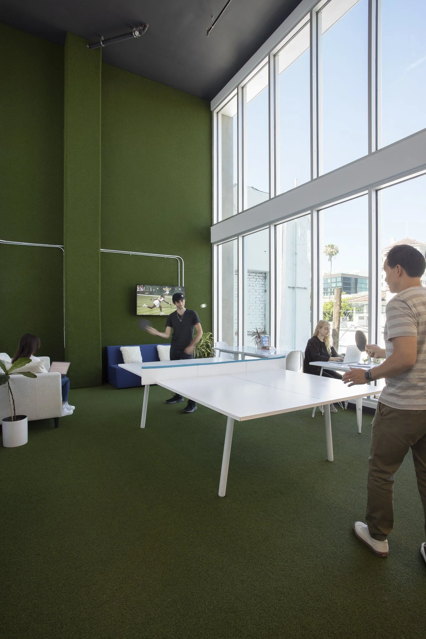 An indoor lounge area with green carpet and walls, large windows allowing natural light, with people engaging in leisure activities, including a person playing table tennis, a woman playing on a laptop, and another woman sitting on a sofa.