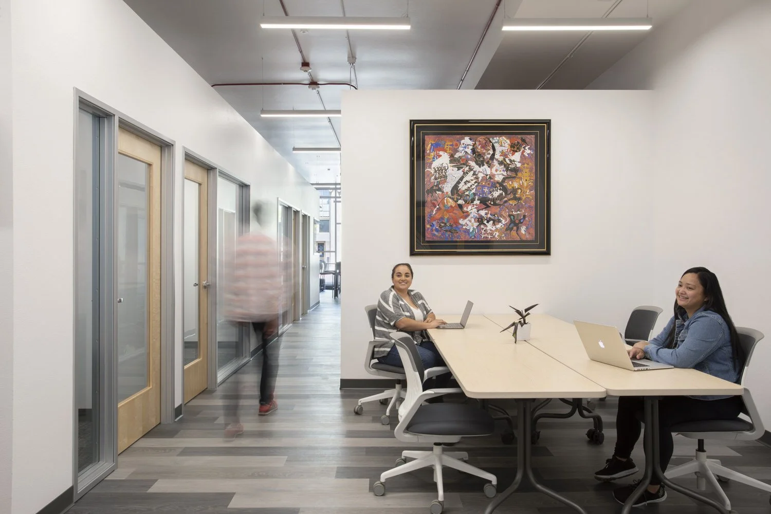 Two women sitting at a conference table with laptops in a modern office, one smiling at the camera, with a colorful abstract painting on the wall behind them.