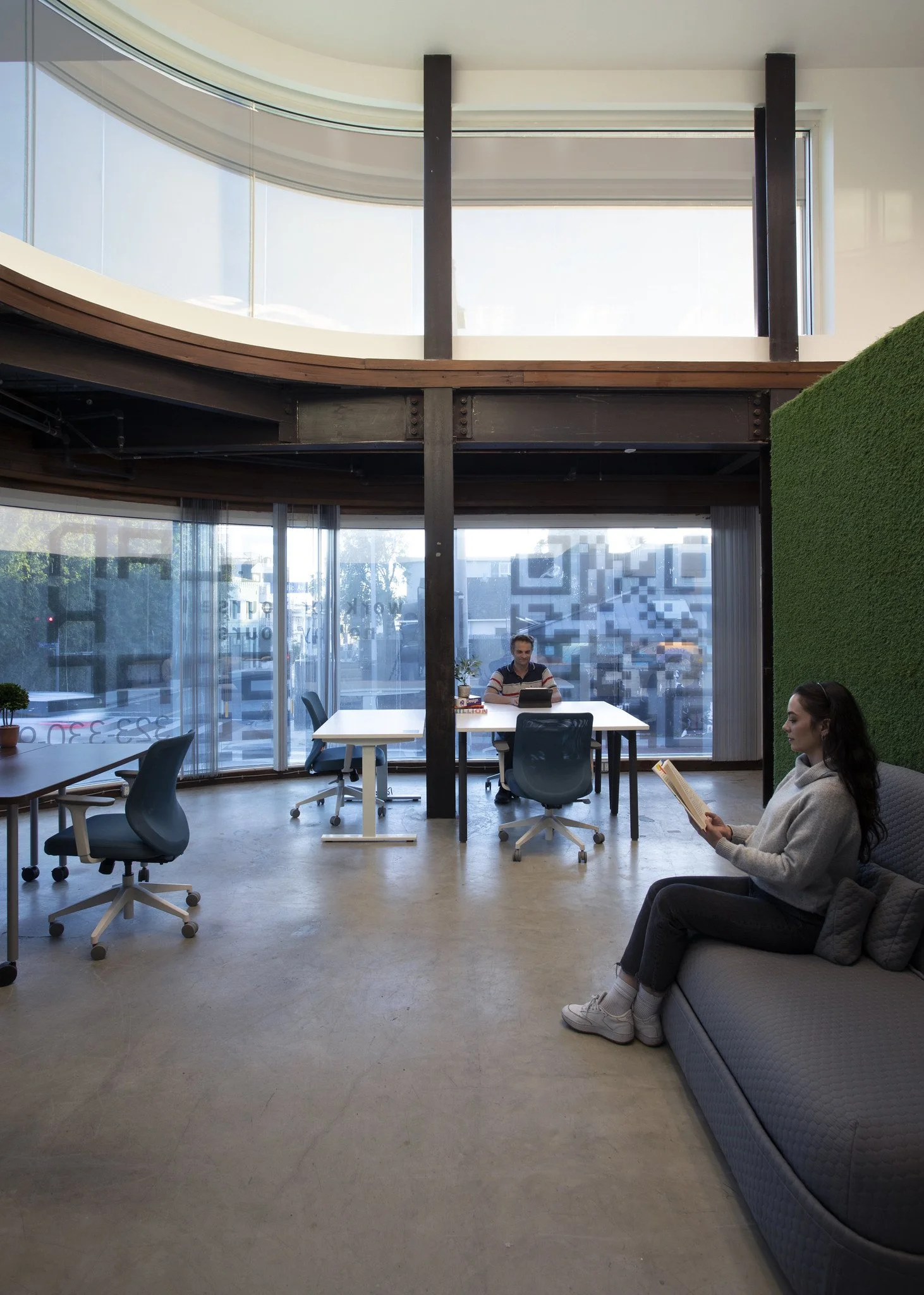 An interior office space with a large curved window, modern chairs, a couch with a woman reading, and a man working at a desk in the background.