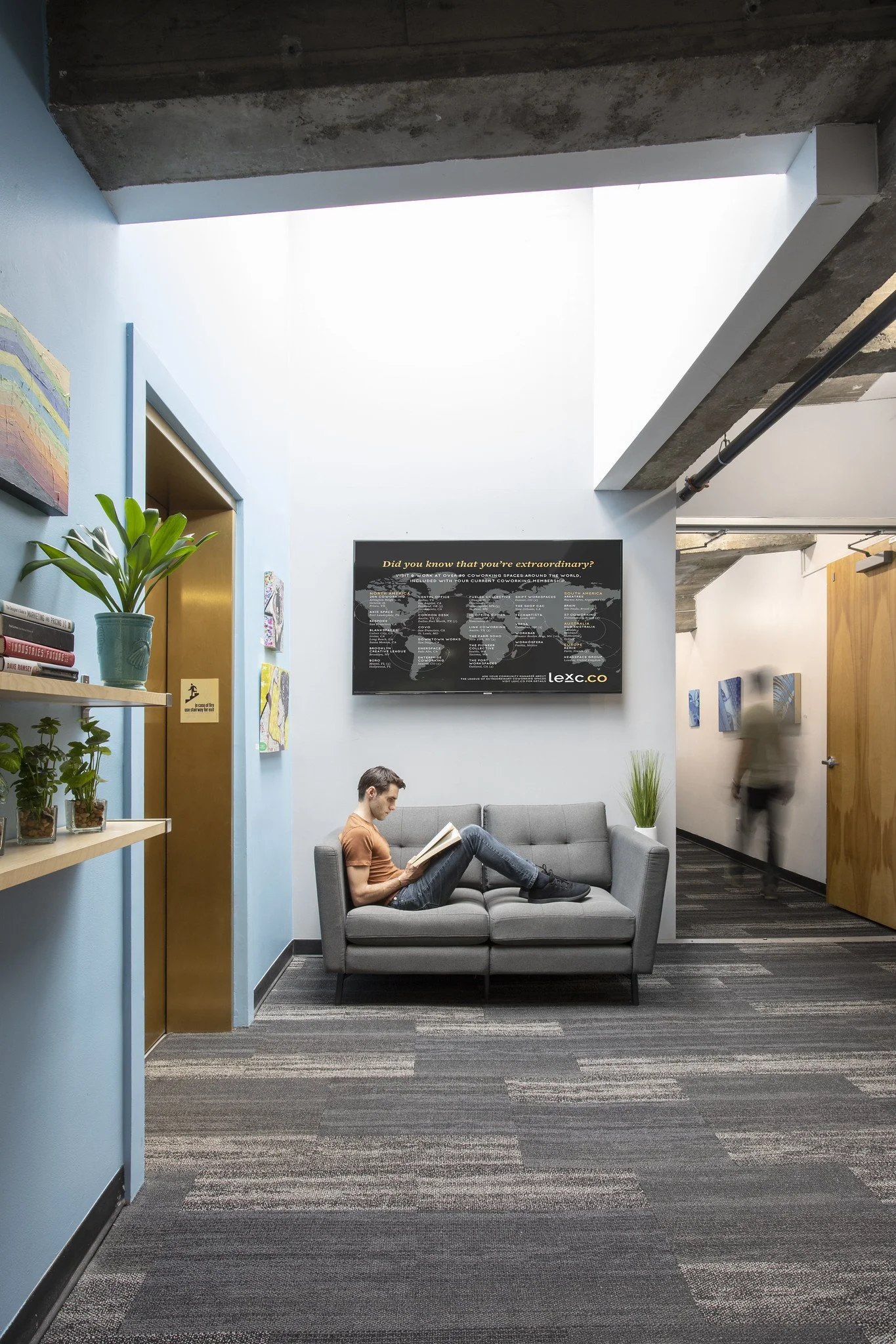 A man sitting on a gray couch in a modern office lobby reading a book, with a TV screen displaying a map on the wall behind him, light blue walls, plants, and a door to an elevator.