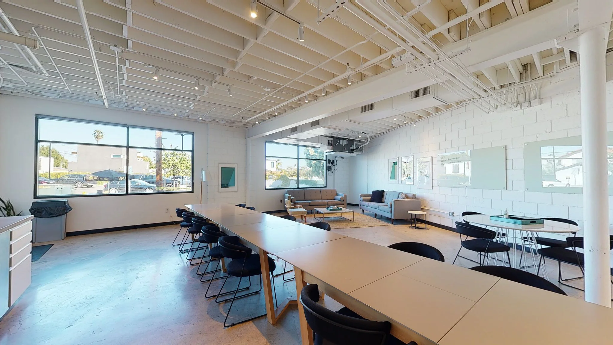 A spacious, modern office or coworking space with large windows, a long table with multiple chairs, a seating area with sofas, artwork on the white brick walls, and a high ceiling with exposed pipes and beams.