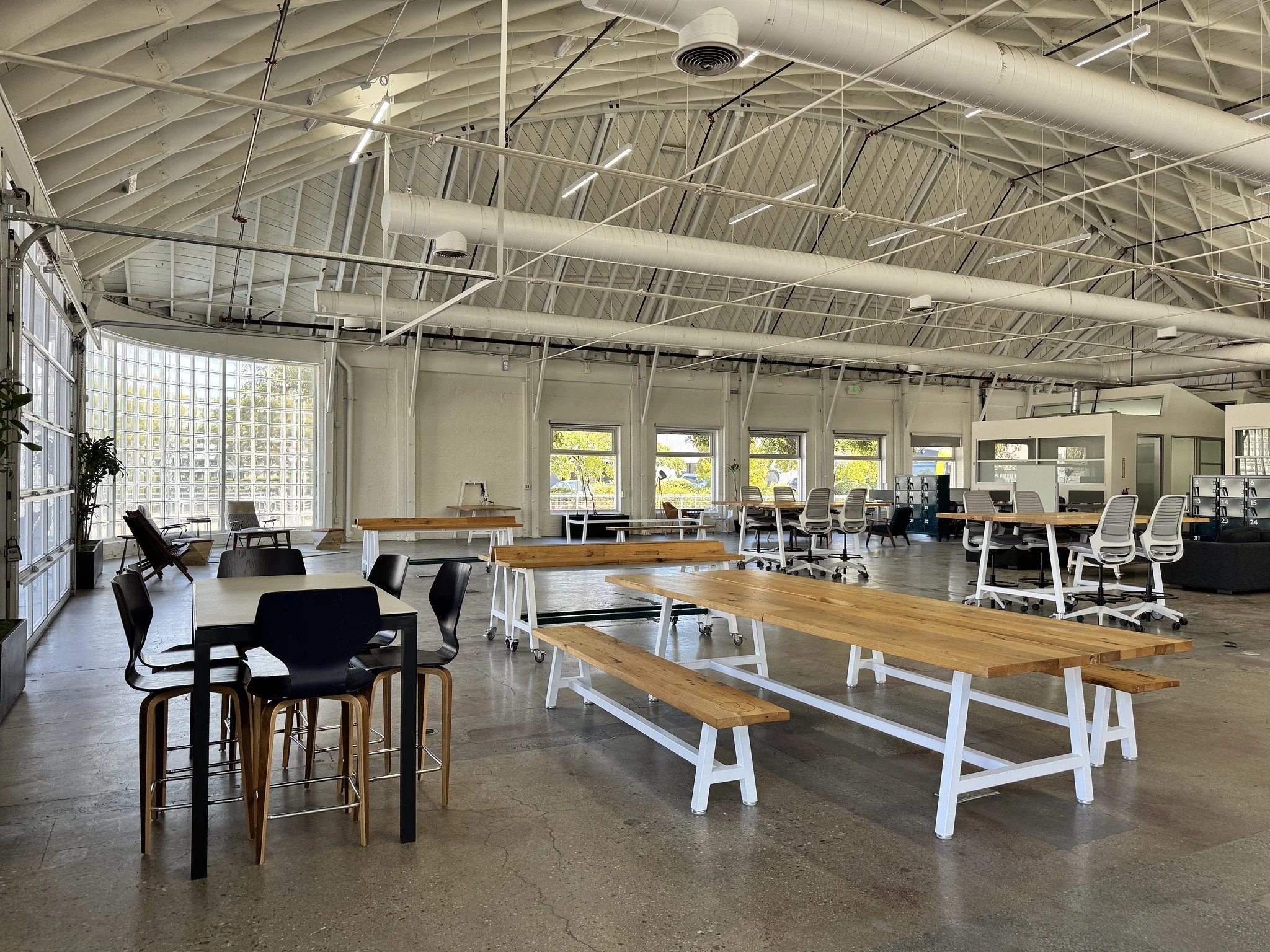 Modern open-plan coworking space at Blankspaces, CA, with long wooden tables, white chairs, a dining table with black chairs, lounge chairs, and large windows allowing natural light.