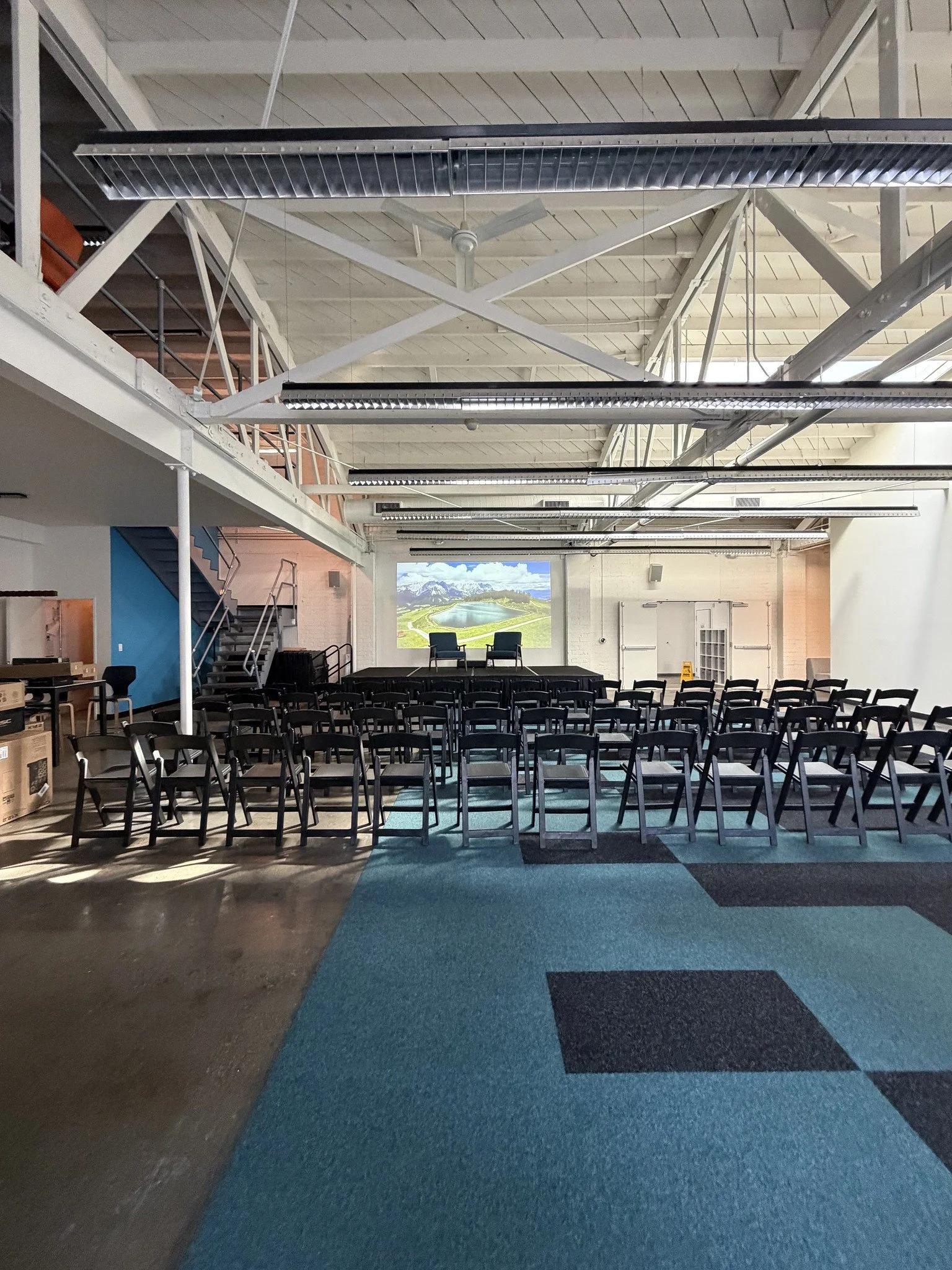 An empty indoor event space with rows of black chairs facing a stage with two chairs and a large screen showing a mountain lake scene.