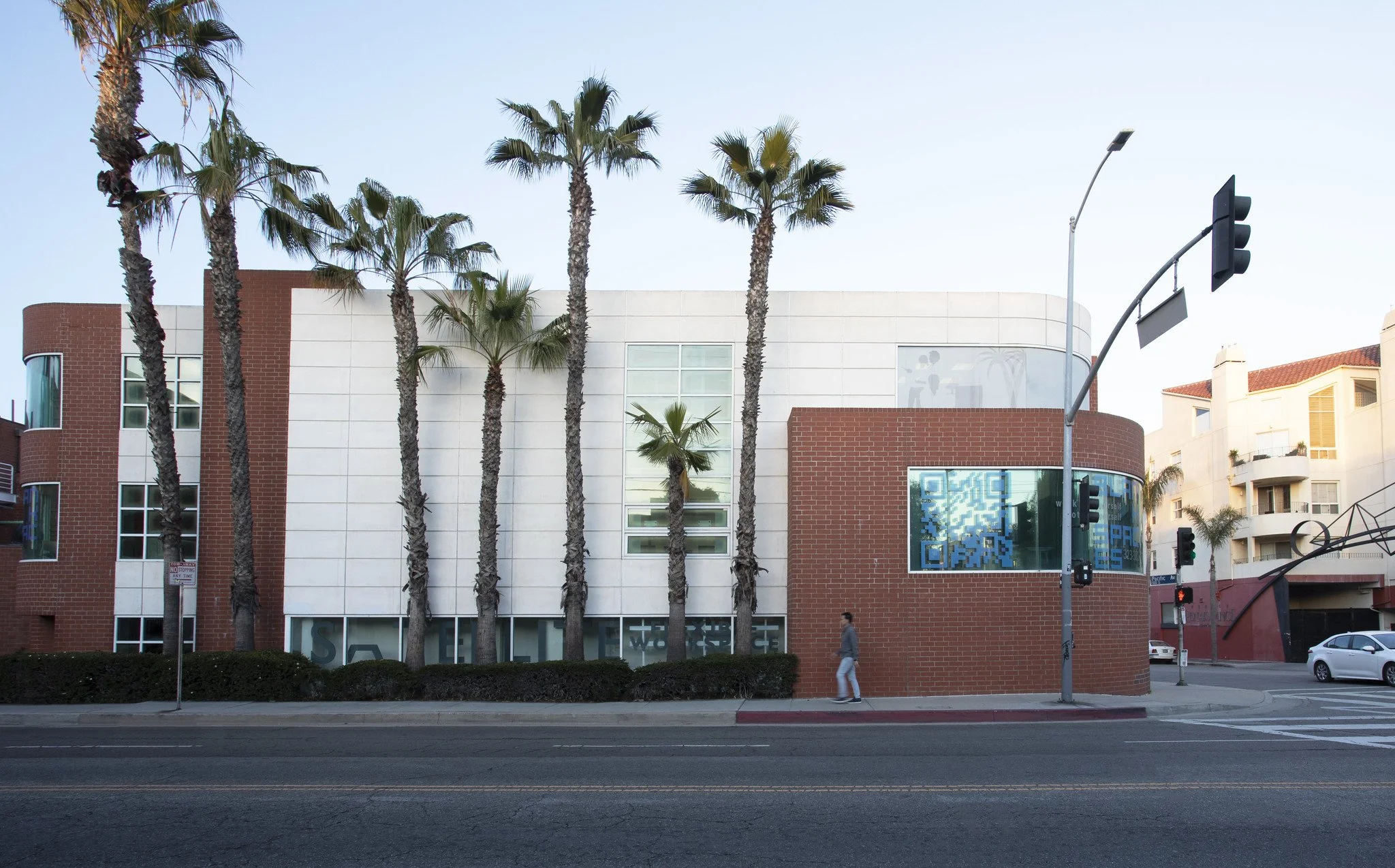 A modern building with white and red brick exterior, seven tall palm trees in front, and a woman walking past on the sidewalk.