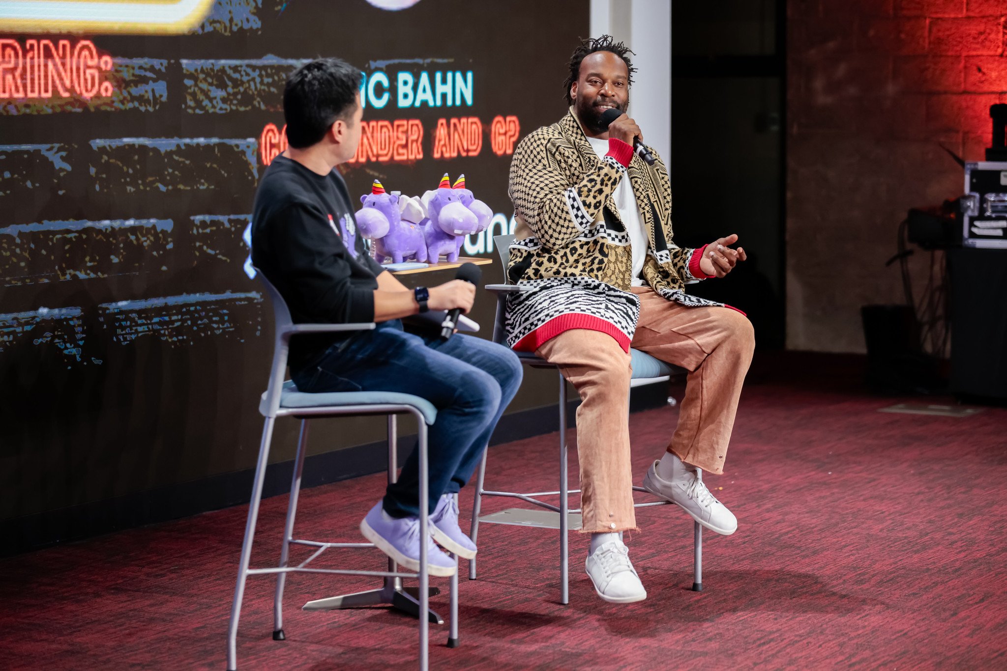 Two men sitting on stage chairs having a conversation, one holding a microphone, with a screen in the background displaying names and graphics, and plush unicorn toys on a table behind them.